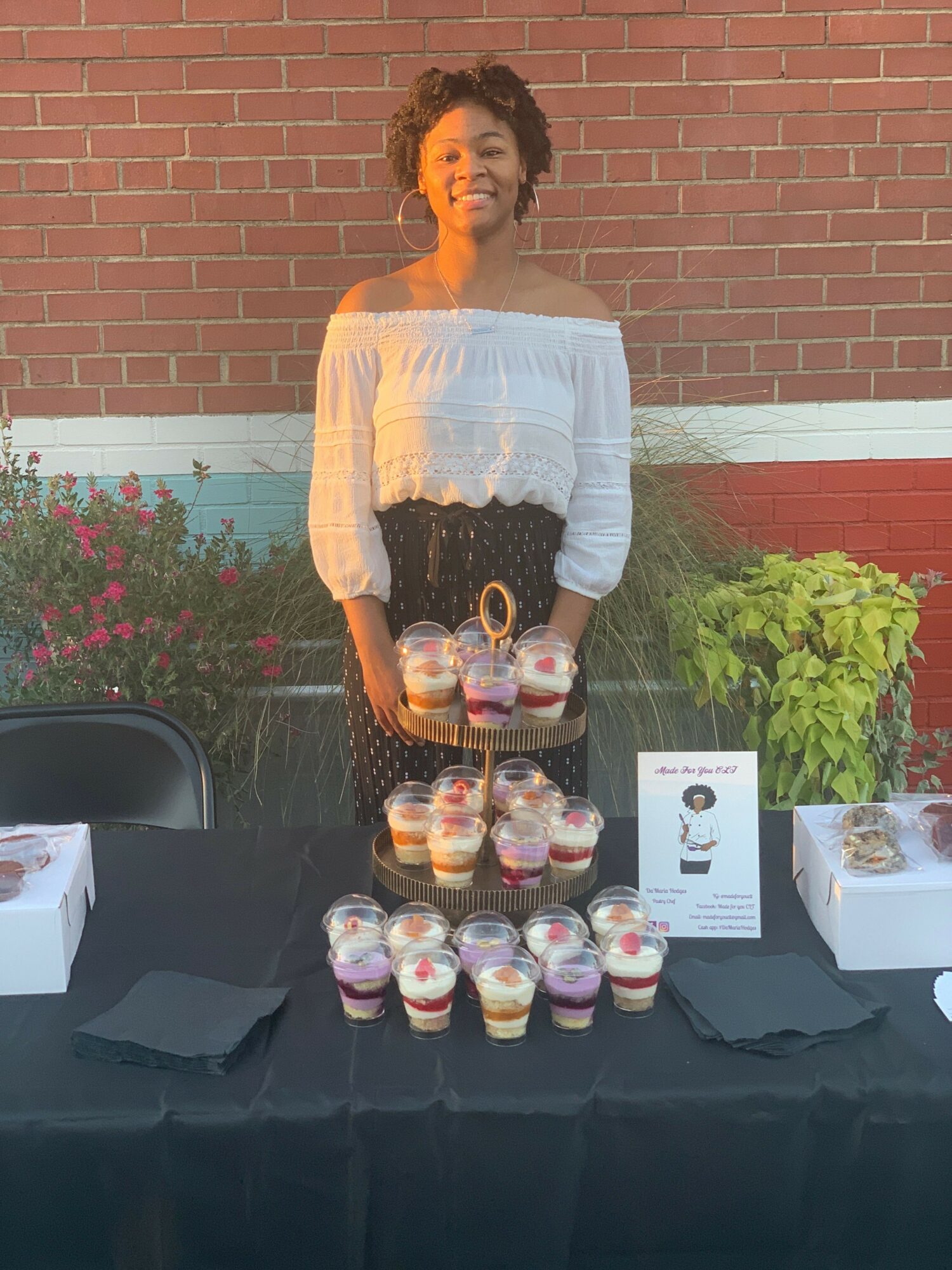 Woman standing behind a table with jars and a sign, brick wall background, plants nearby.