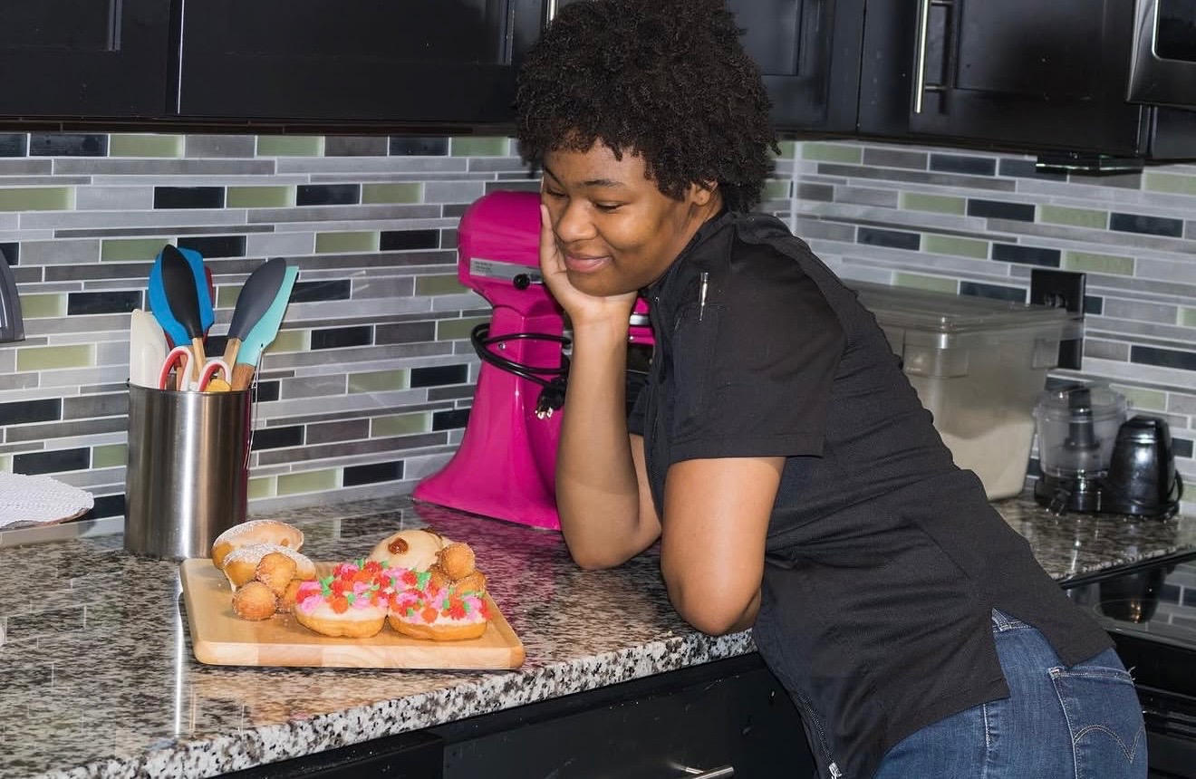 Child with curly hair leaning on a kitchen counter, looking at a tray of decorated cupcakes, with utensils nearby.