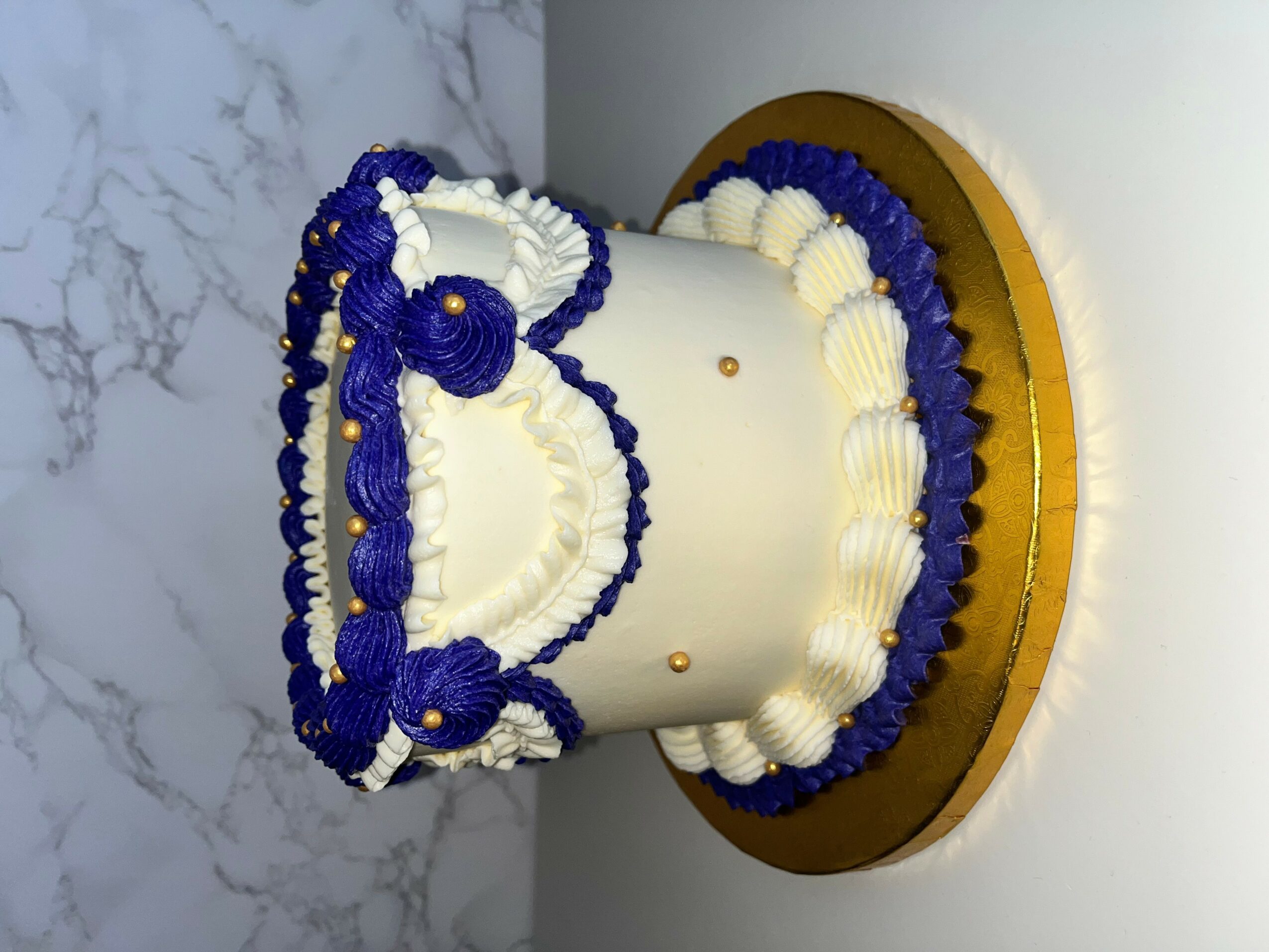 A white cylindrical cake decorated with blue and white icing, placed on a round gold-colored base.