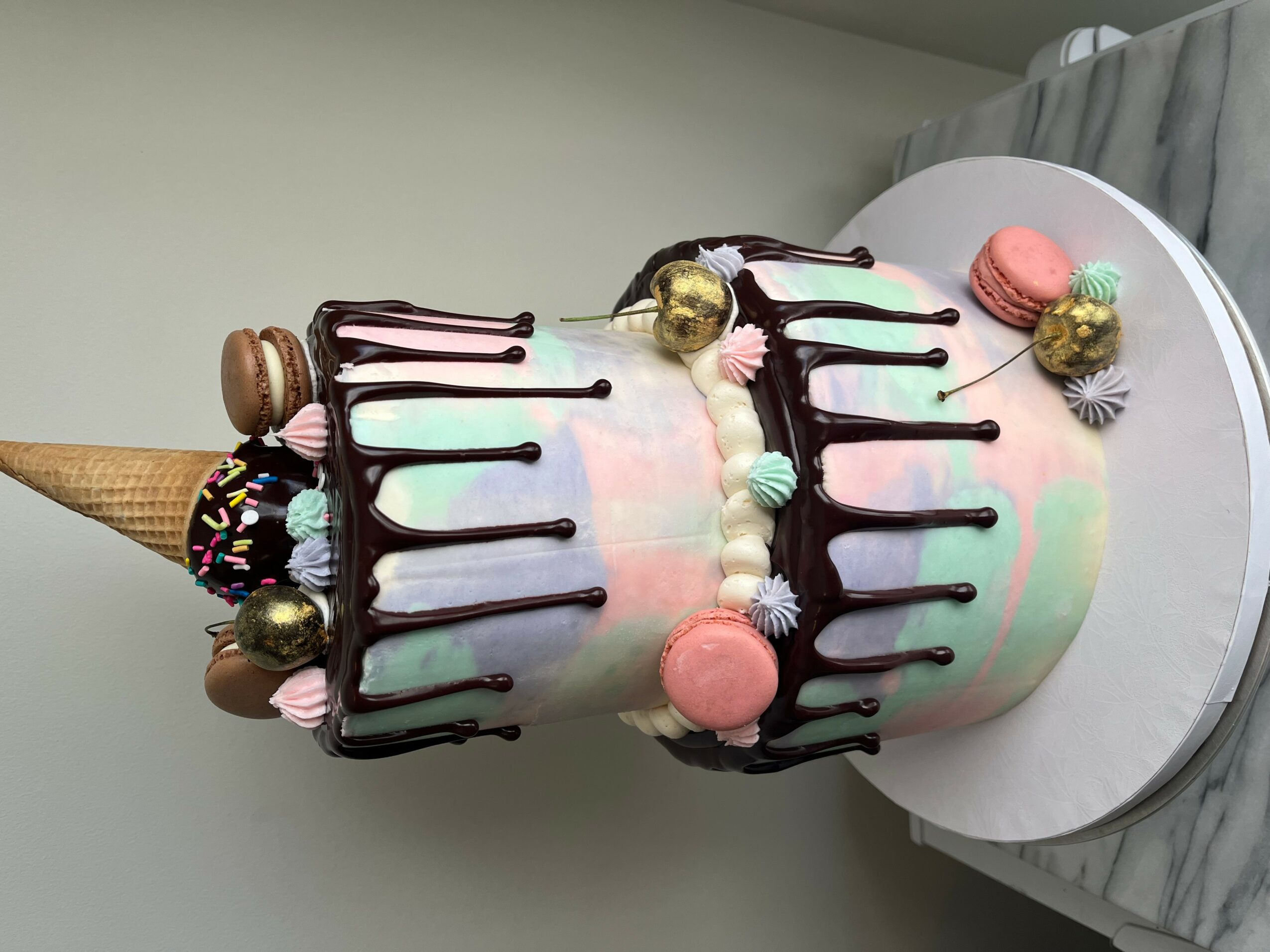 Colorful layered cake decorated with macarons, candies, and chocolate drizzle, with an ice cream cone on top, on a white surface.