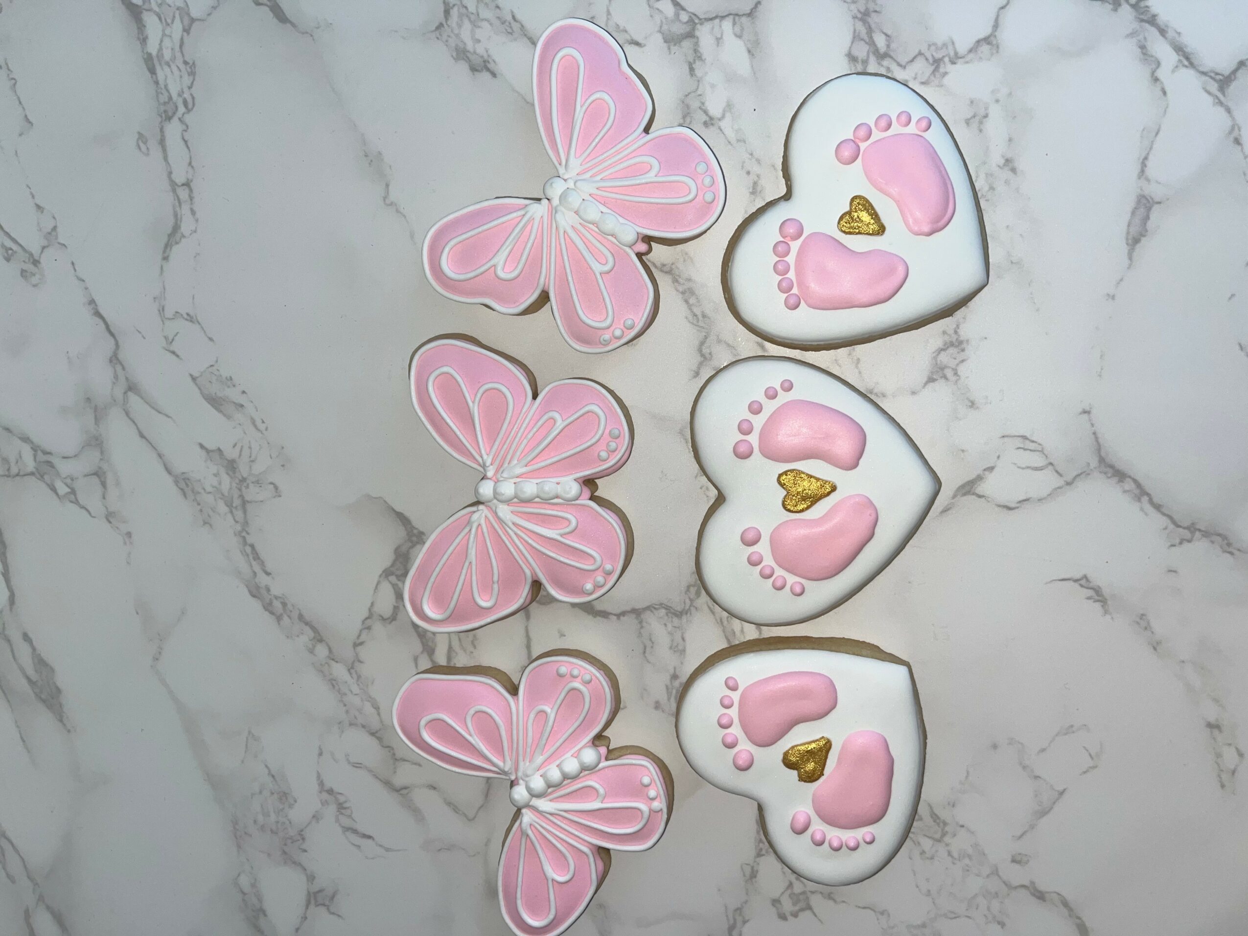Three pink butterfly cookies and three white heart-shaped cookies with pink and gold decorations on a marble surface.