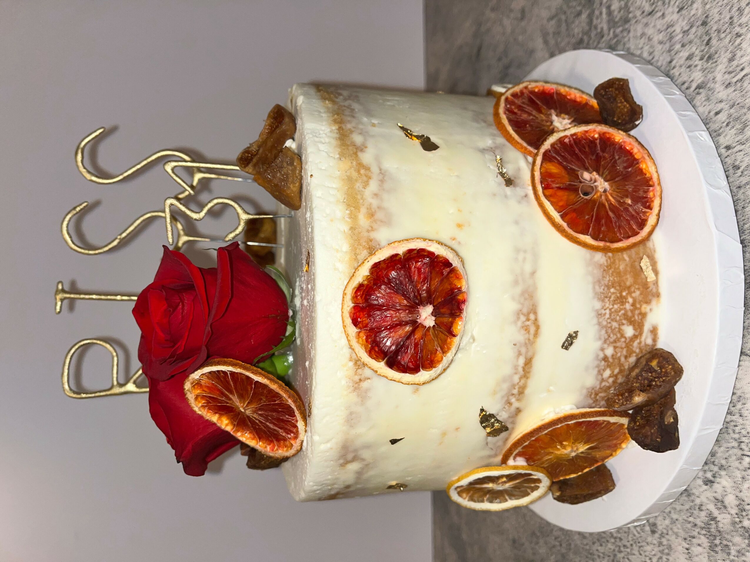 Round cake decorated with dried orange slices, a red flower, and small brown decorations, on a white plate.