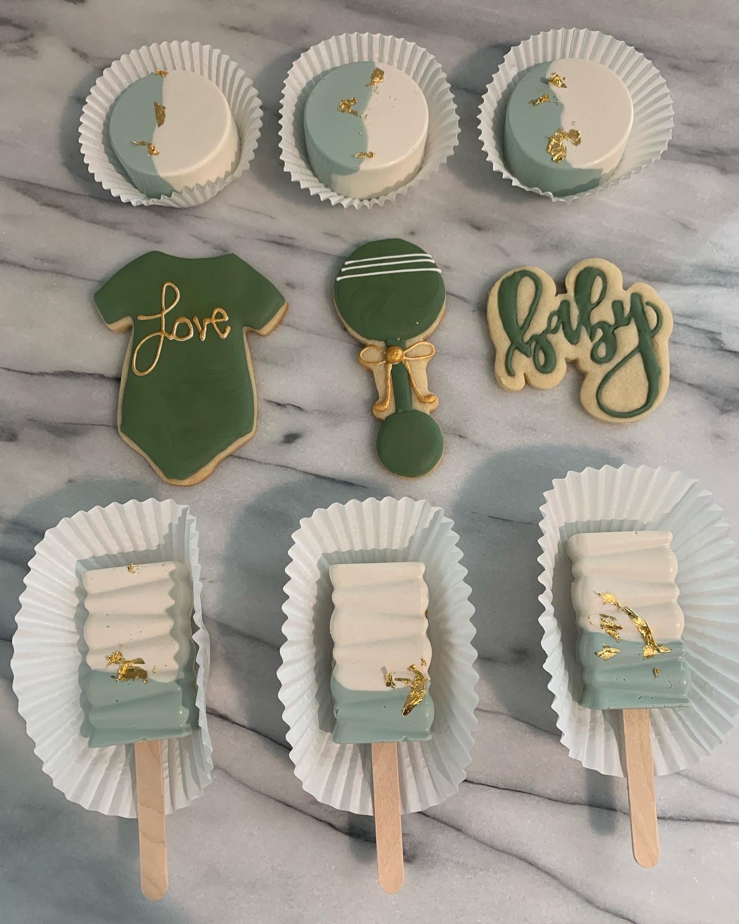 Decorative cookies on a marble surface, including a love shirt, golf ball, golf club, and three cookies with blue and white designs, each in paper cups.