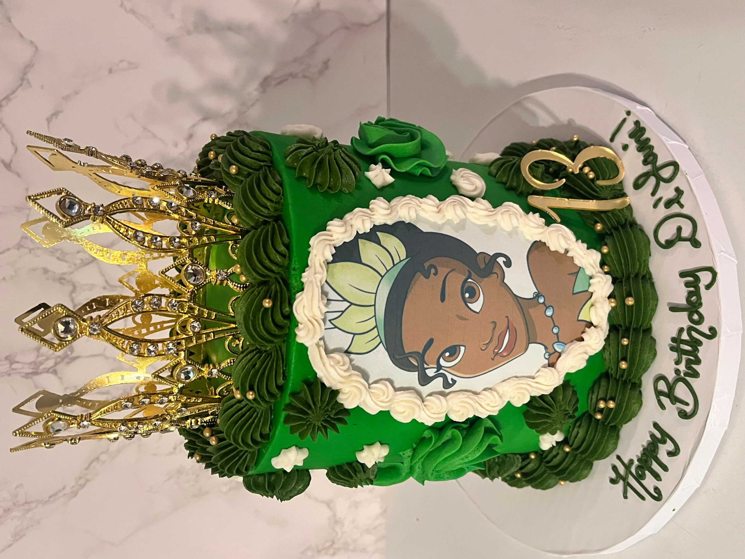 Decorative birthday cake with a princess illustration, green and white frosting, and gold crown decorations, on a white surface.