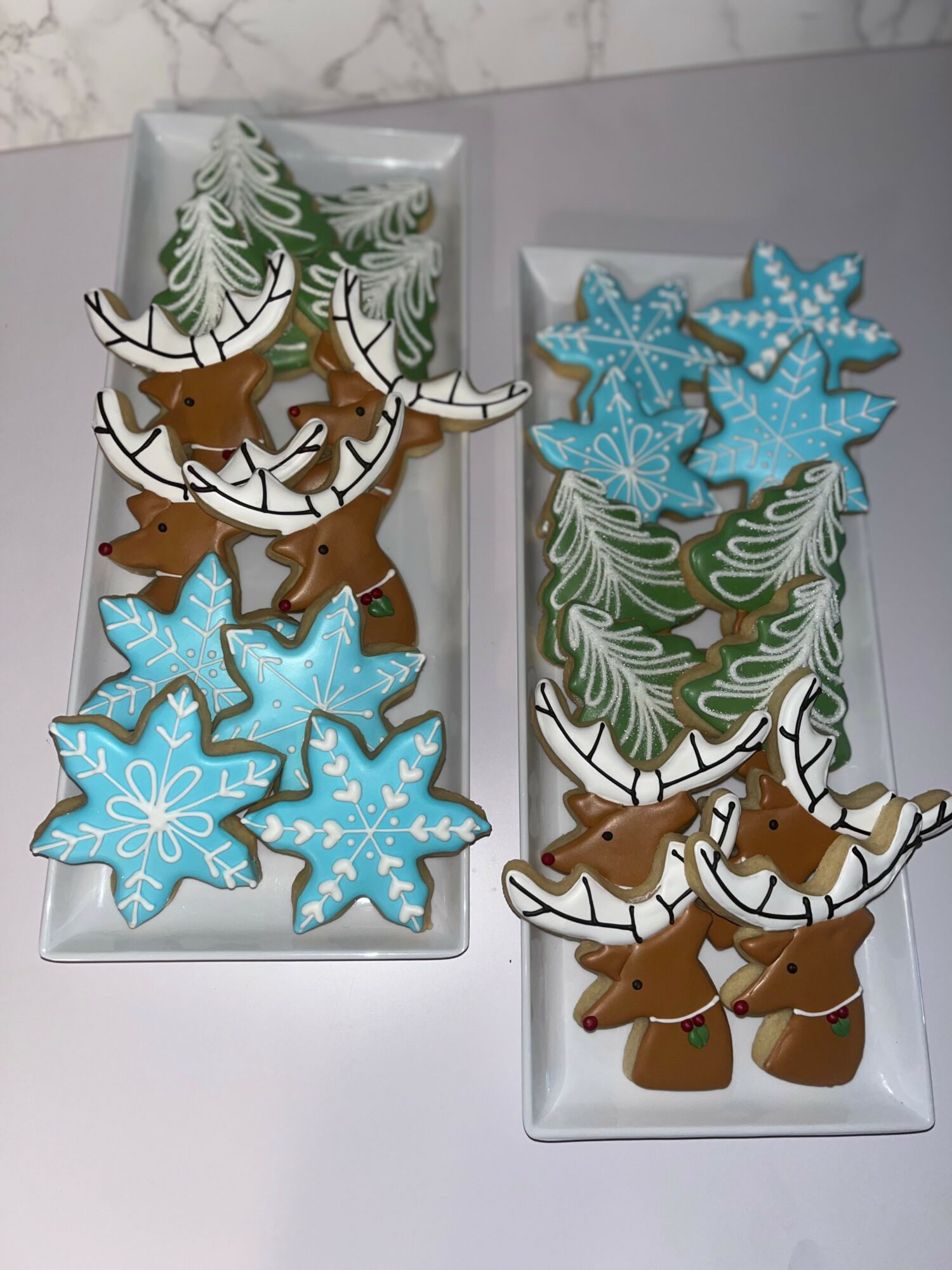 Two trays of decorated cookies, including reindeer and snowflakes, with green and white icing, on a marble surface.
