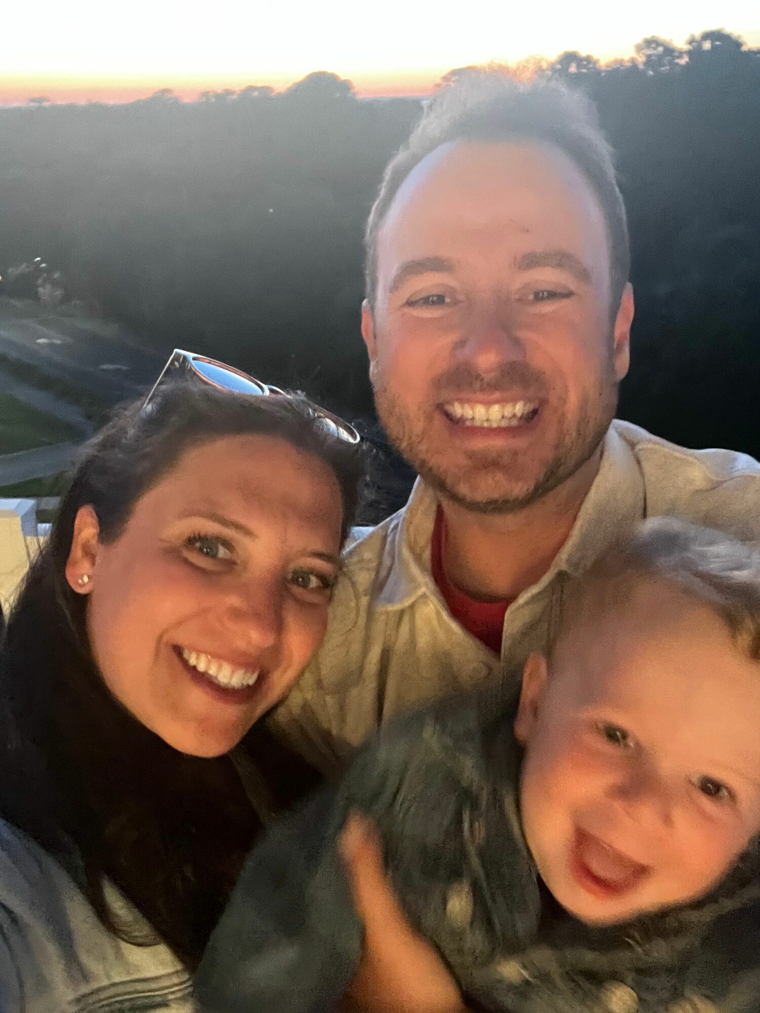 Three people smiling outdoors during sunset, with a child and two adults close together.