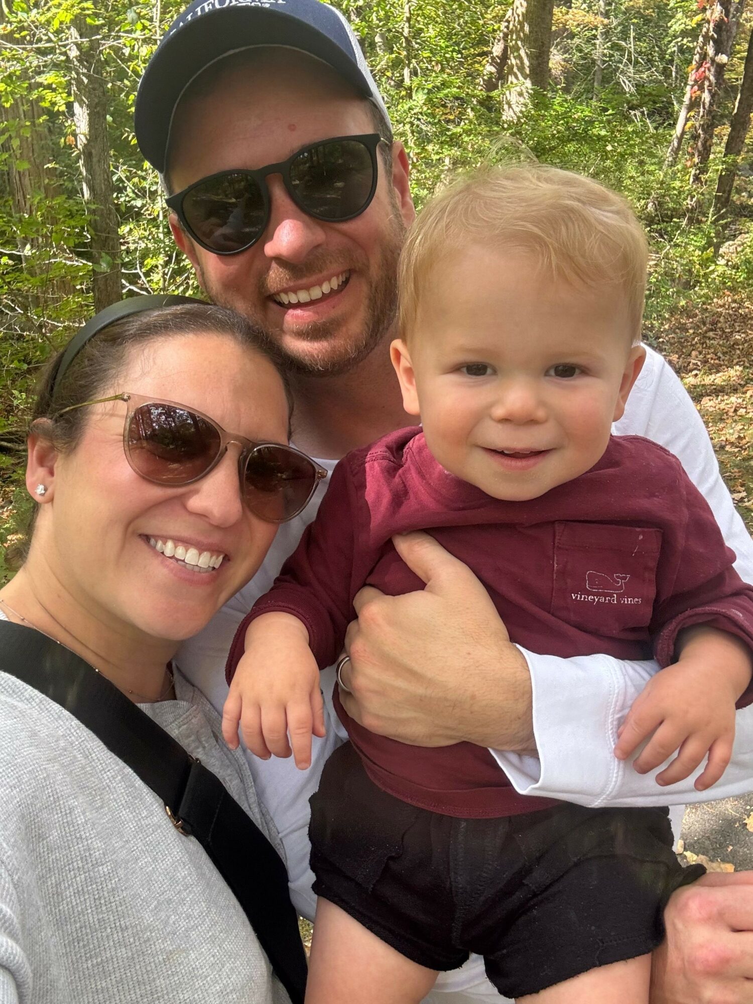 Two smiling women and a young child outdoors in a wooded area, with trees and sunlight visible in the background.