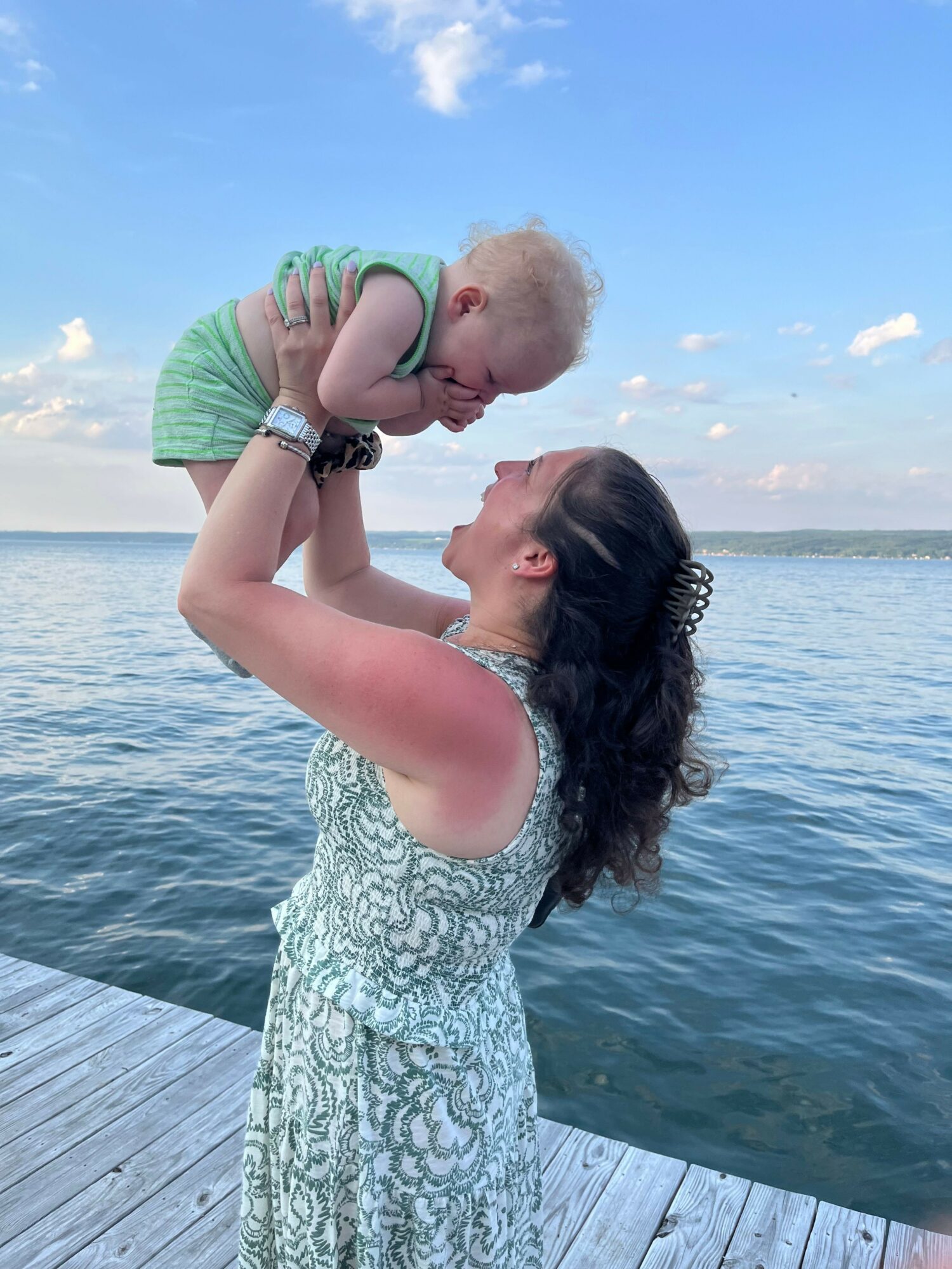 Woman holding a baby near water on a dock, smiling and touching noses, sky with clouds in background.