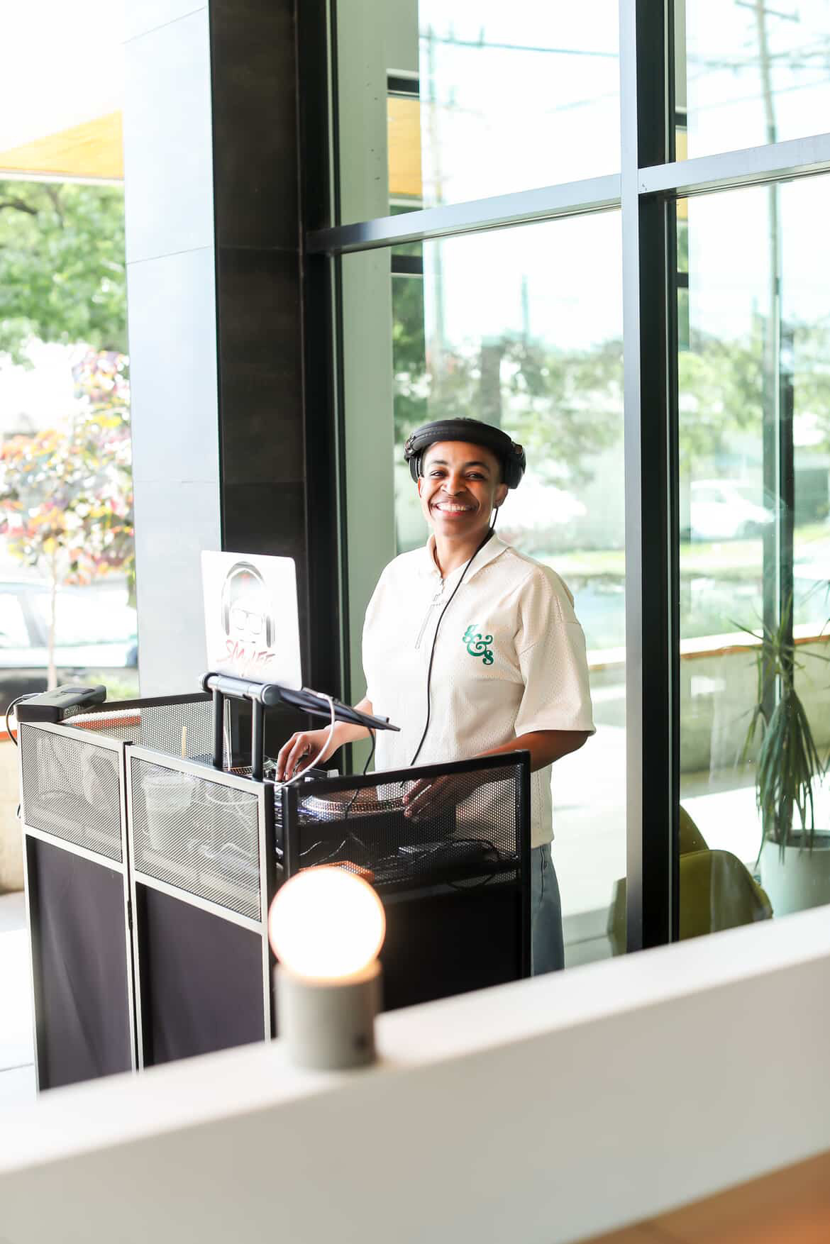 Smiling person standing at a reception desk inside a building with large glass windows, natural light, and greenery outside.