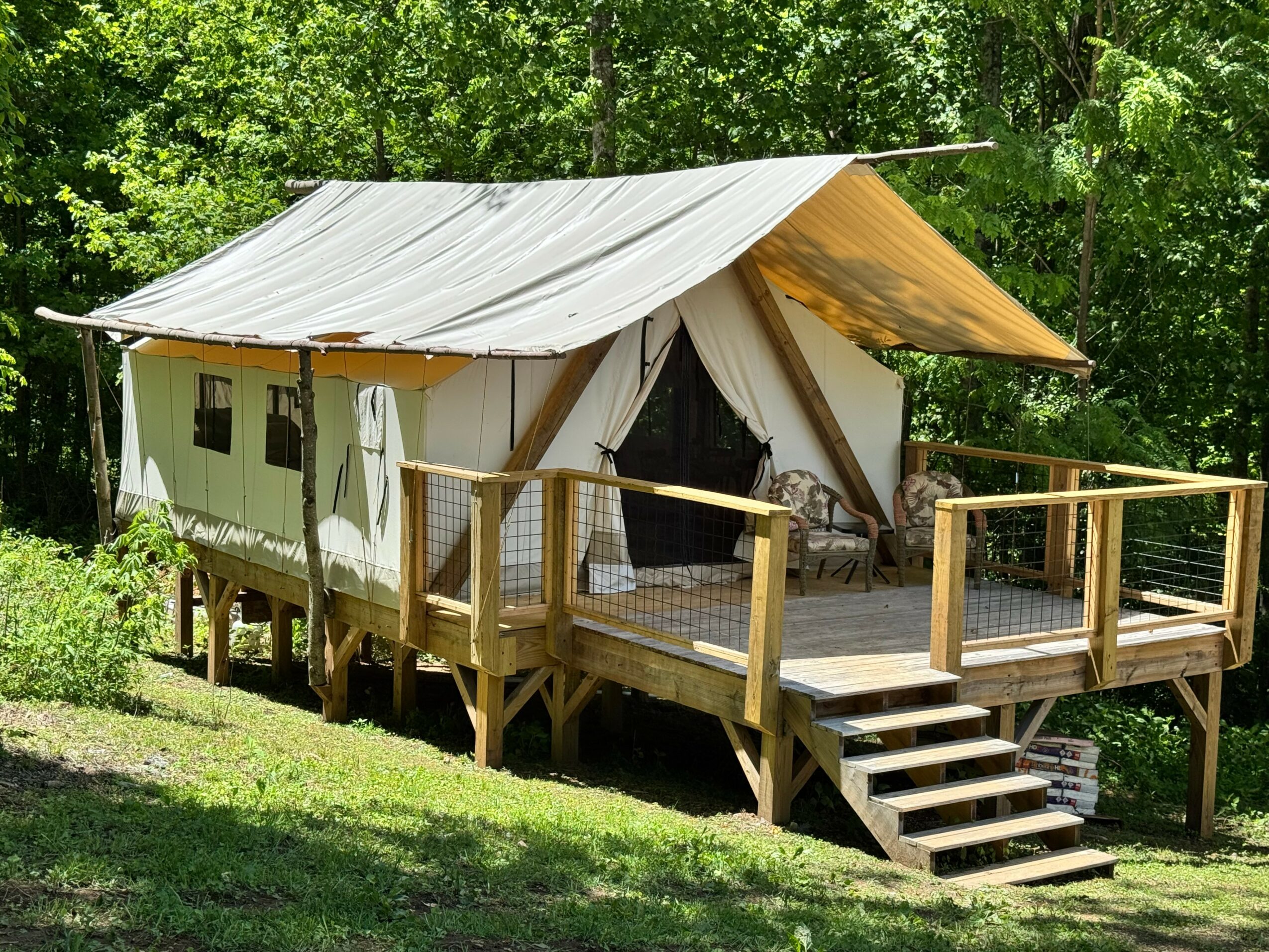 Wooden cabin with a raised deck, surrounded by green trees, with a sloped roof and an open front entrance.