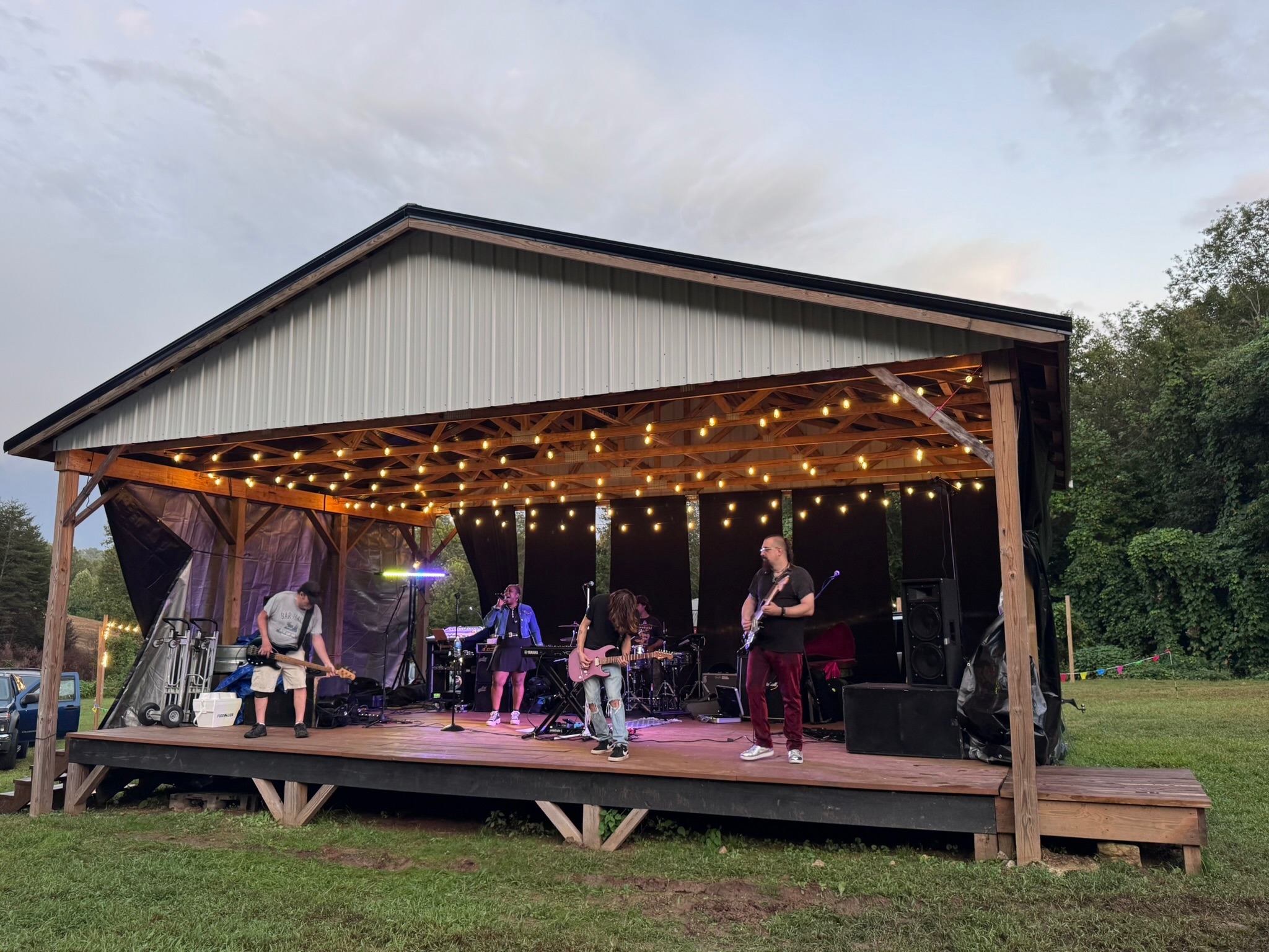 Band performs on an outdoor stage with string lights, surrounded by trees, during evening.