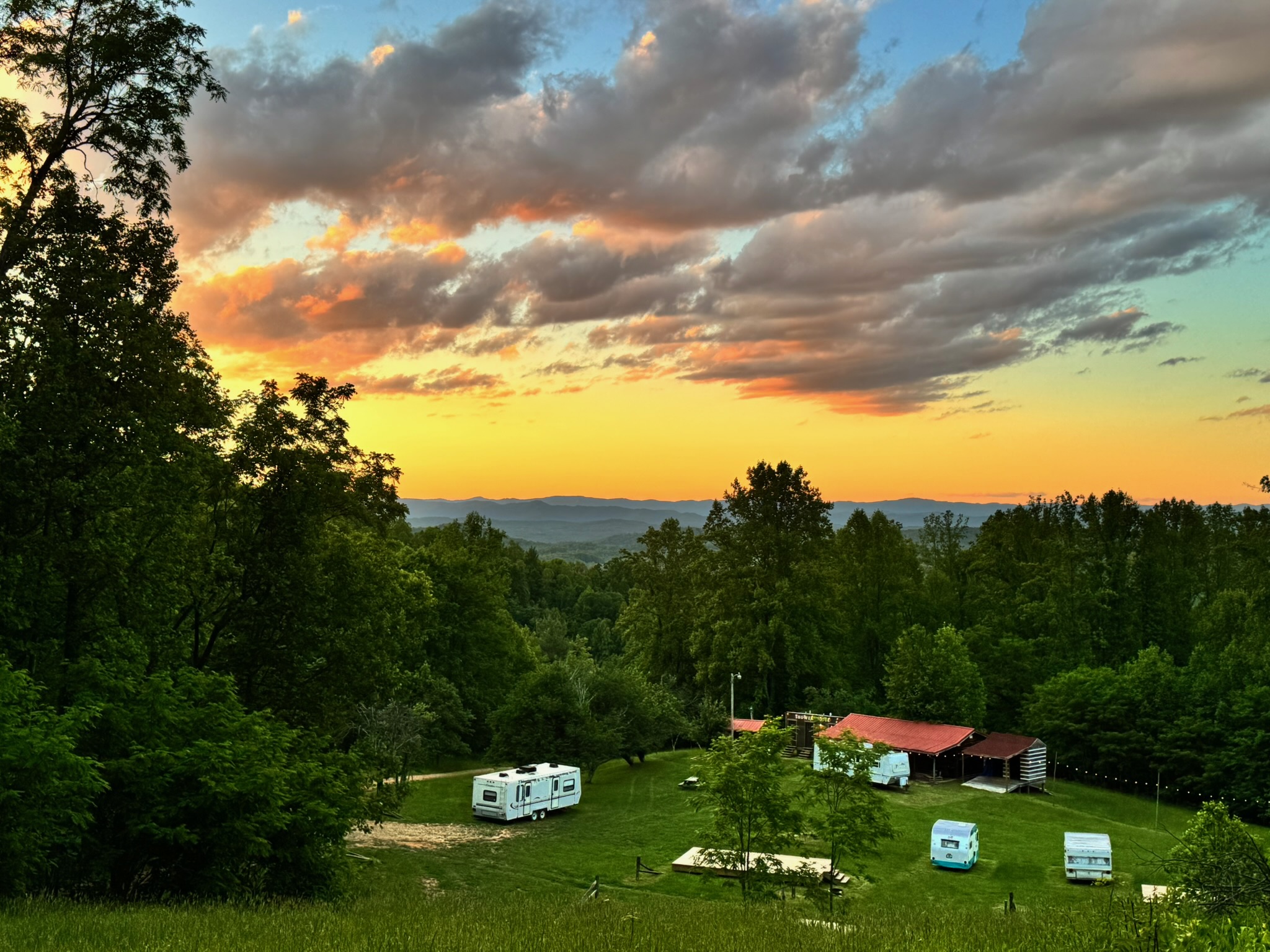 Sunset sky with clouds over a green landscape, trees, and parked trailers and caravans, with a red-roofed building in the background.