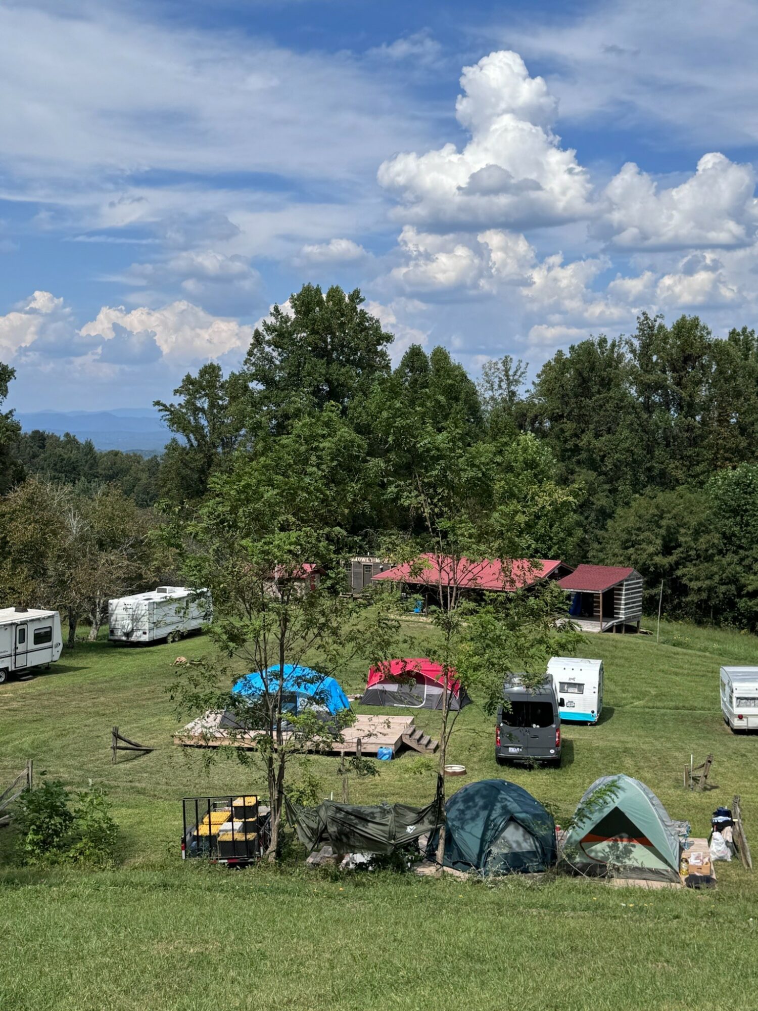 Campsite with tents, RVs, and trees under a partly cloudy sky, surrounded by greenery and open space.
