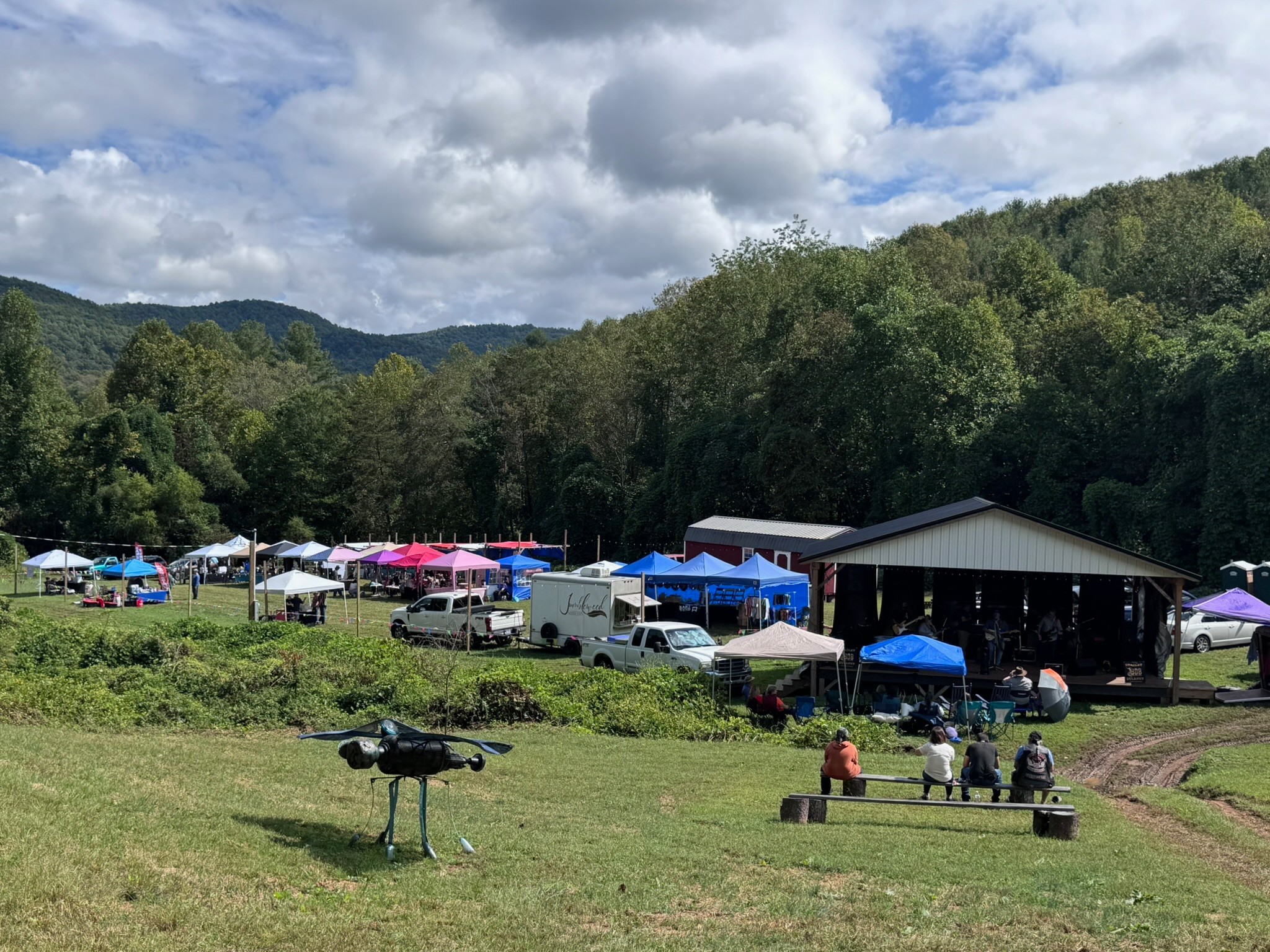 Outdoor event with tents, vehicles, and people in a grassy area surrounded by trees and hills under a partly cloudy sky.