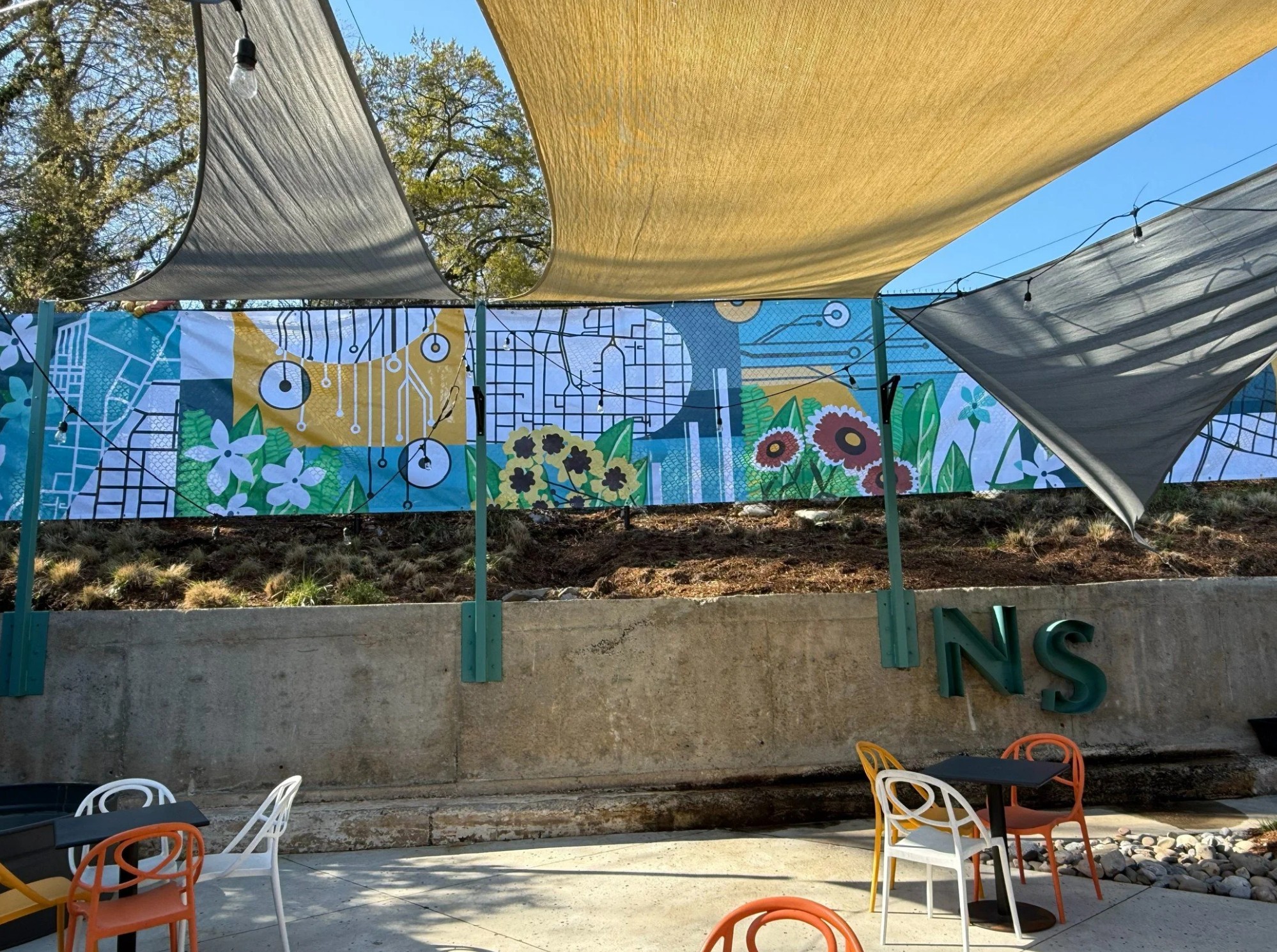 Colorful mural with flowers and bees, shaded outdoor seating area with chairs, and fabric canopies overhead.