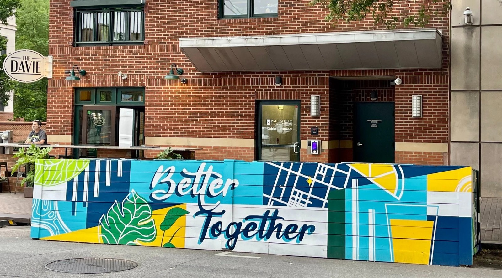 Colorful mural with cityscape and text 'Better Together' on a sidewalk barrier in front of a brick building.