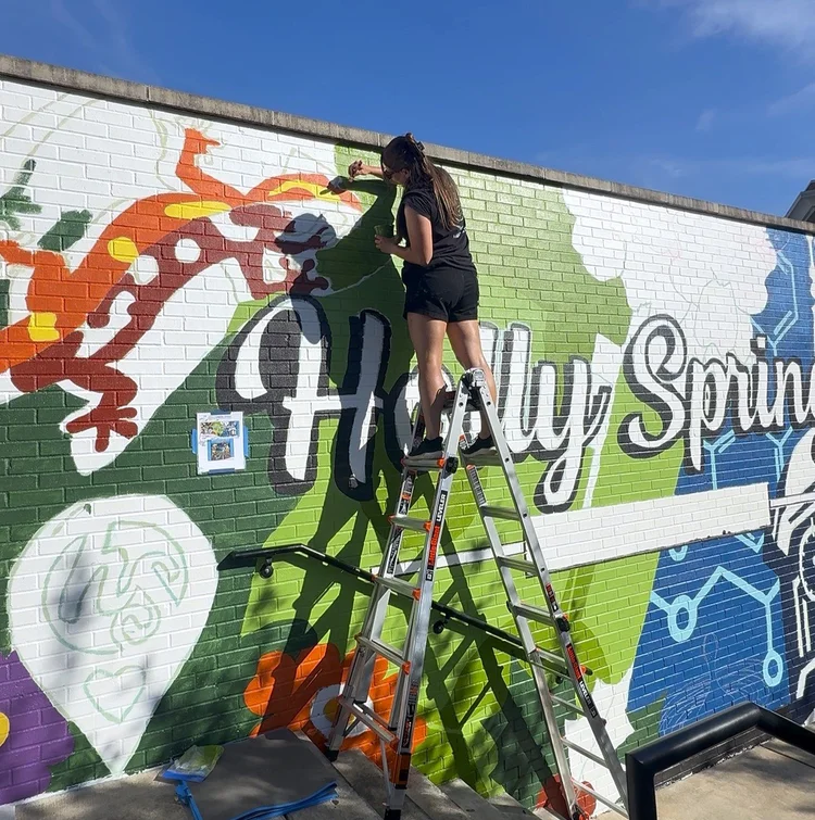 Person painting a colorful mural on a brick wall, standing on a ladder outdoors under a blue sky.