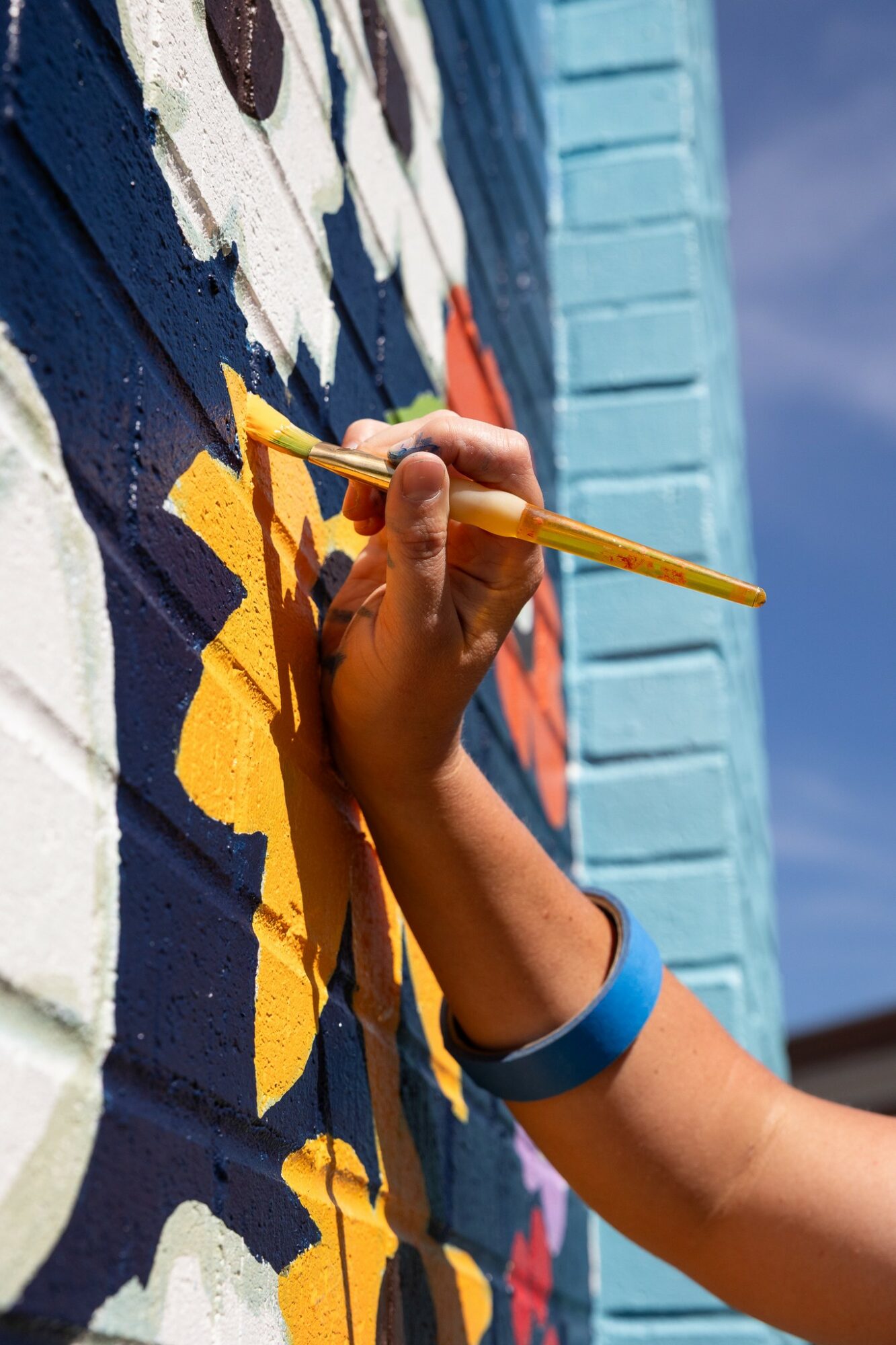 Person painting a colorful mural on a wall with a yellow paintbrush, wearing a blue wristband.