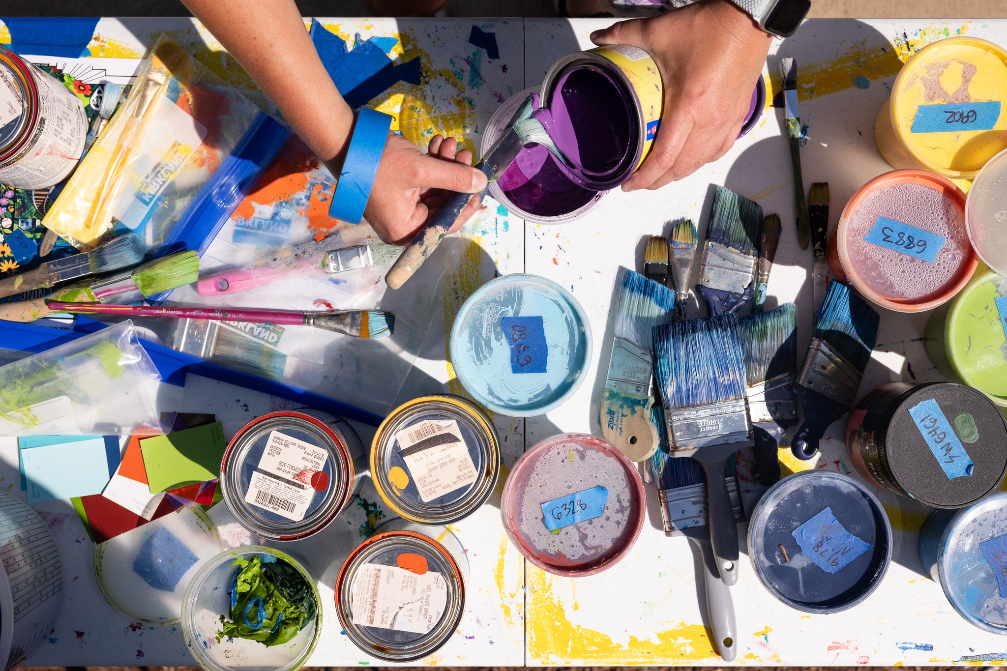 Hands reaching for containers of paint and art supplies on a table, with various colors and tools visible.