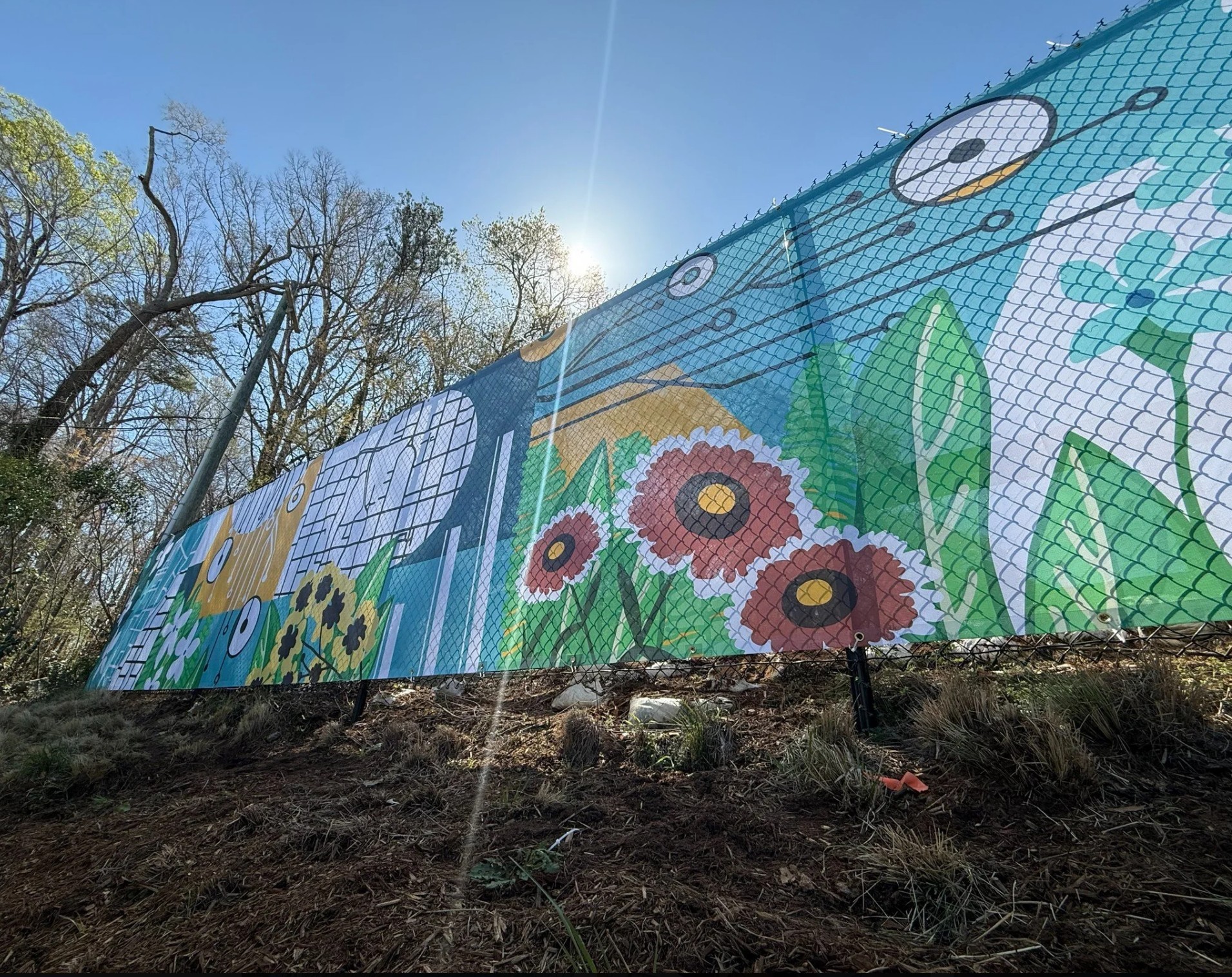 Colorful mural on a building wall with flowers, grass, and a blue sky, viewed from below. Bright sunlight shines on the scene.