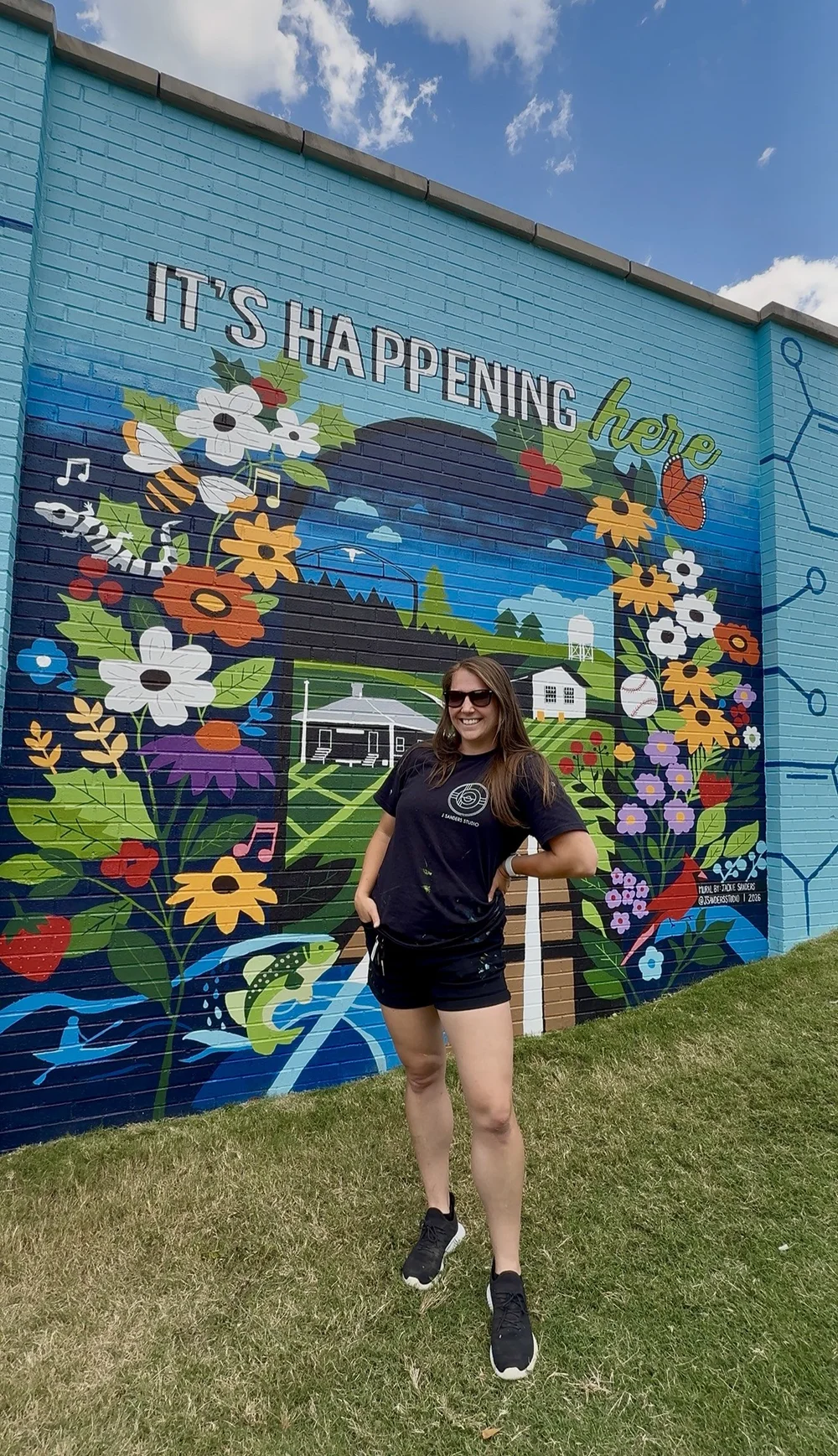 Woman standing in front of a colorful mural with flowers, a rainbow, and the text 'It's happening now'.
