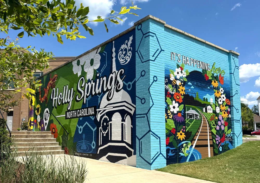 Colorful mural of Holly Springs, North Carolina, with flowers, a train, and a building, on a building wall.