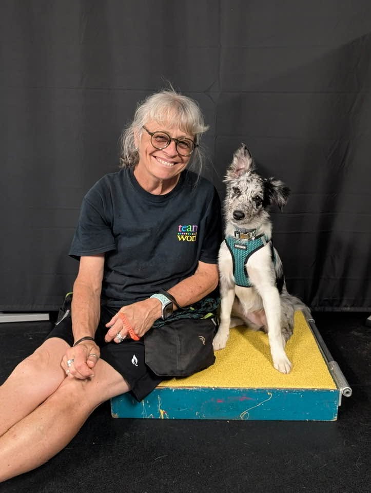 Woman with glasses and gray hair sitting next to a dog on a platform, both smiling, black background.