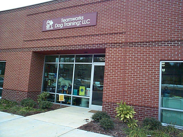 Brick building with a sign that reads 'Teamwork Dog Training LLC' above the entrance, glass door, and windows, with plants outside.