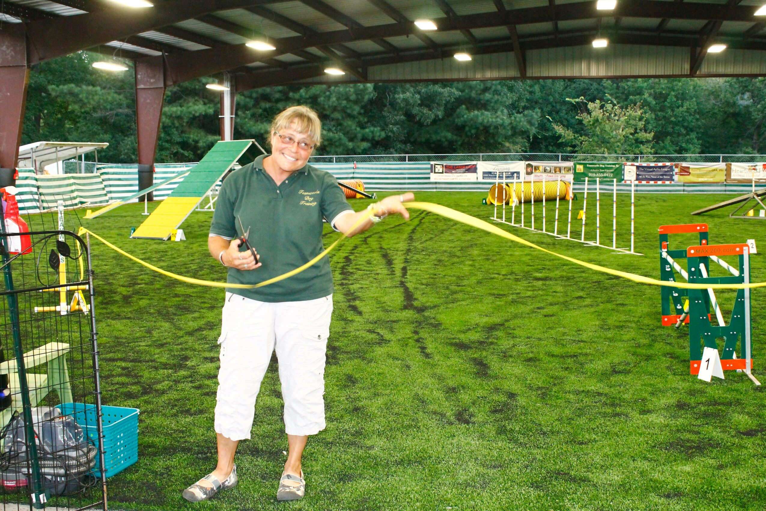 Woman standing on grass field under a roof, holding a yellow rope, with agility equipment in the background.