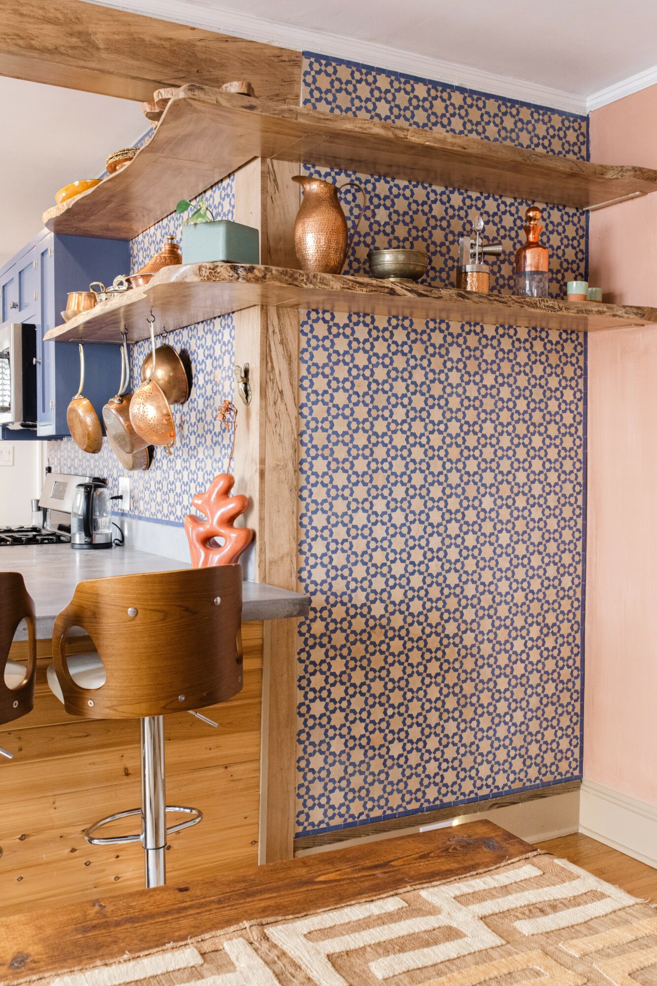 Kitchen with wooden shelves, hanging pots, patterned wall, and a bar stool near a wooden counter.