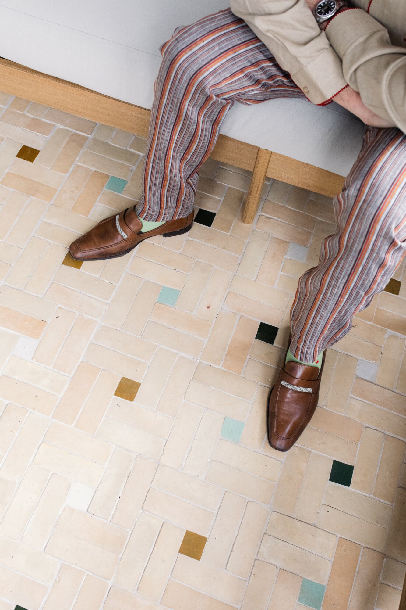 Person sitting on a bench wearing striped pants and brown shoes, with a tiled floor below.