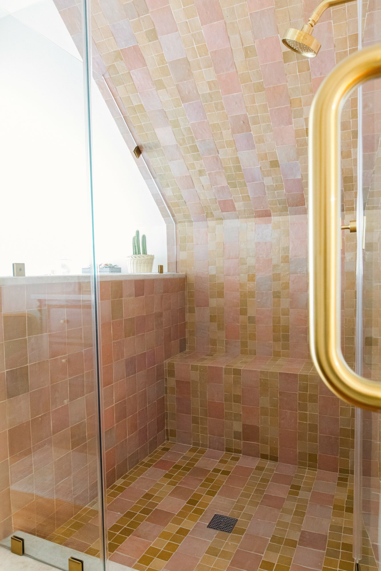 Shower area with pink and yellow tiles, glass door, and a gold handle, featuring a built-in bench and a showerhead.