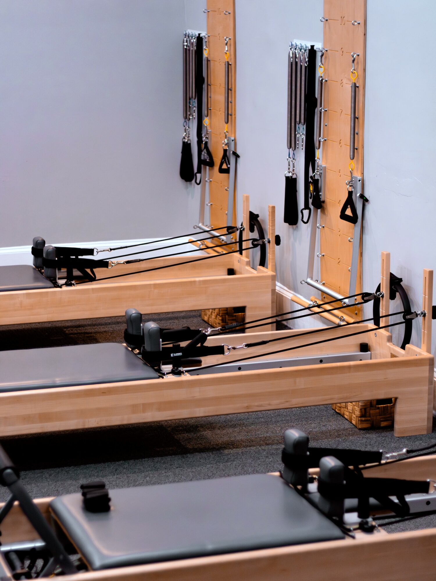 Two wooden Pilates reformer machines with resistance bands and straps in a fitness studio, against a light gray wall.