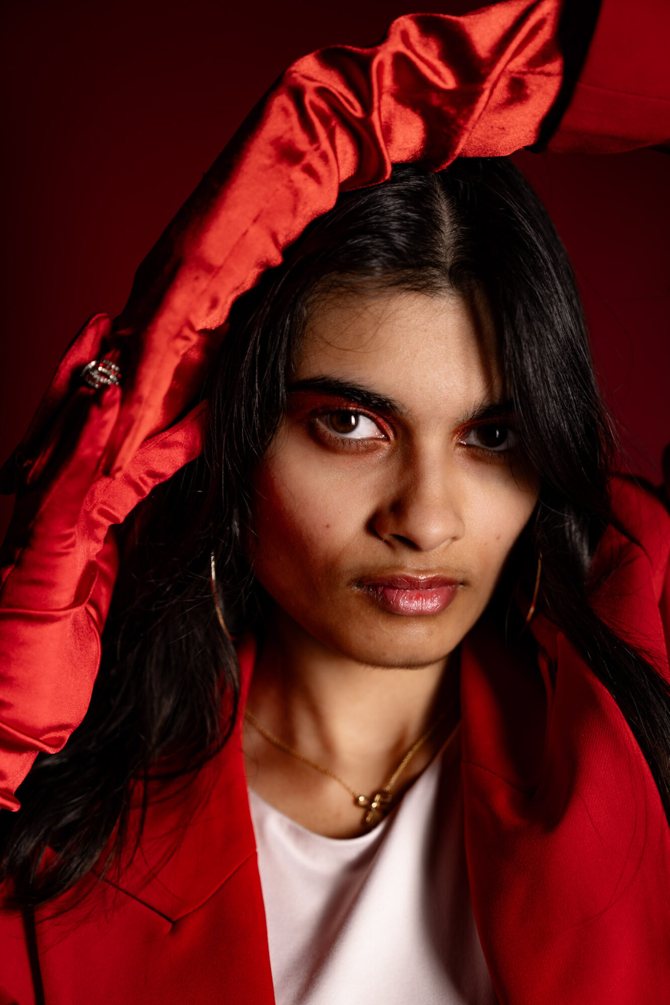 Woman with dark hair and red clothing, holding a red fabric above her head, against a dark background.