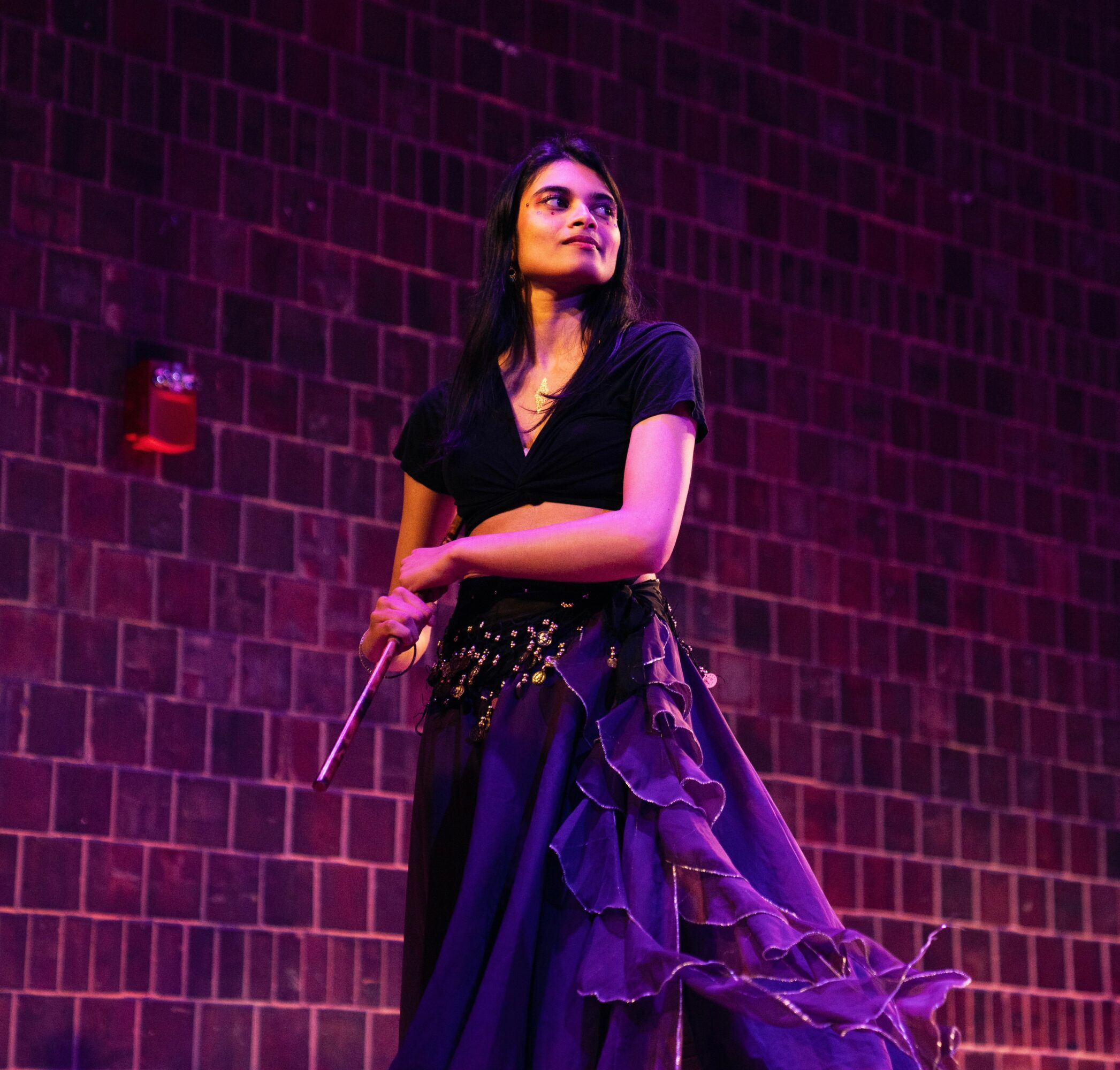 Woman standing against a brick wall, wearing a black top and a flowing skirt, illuminated by purple lighting.
