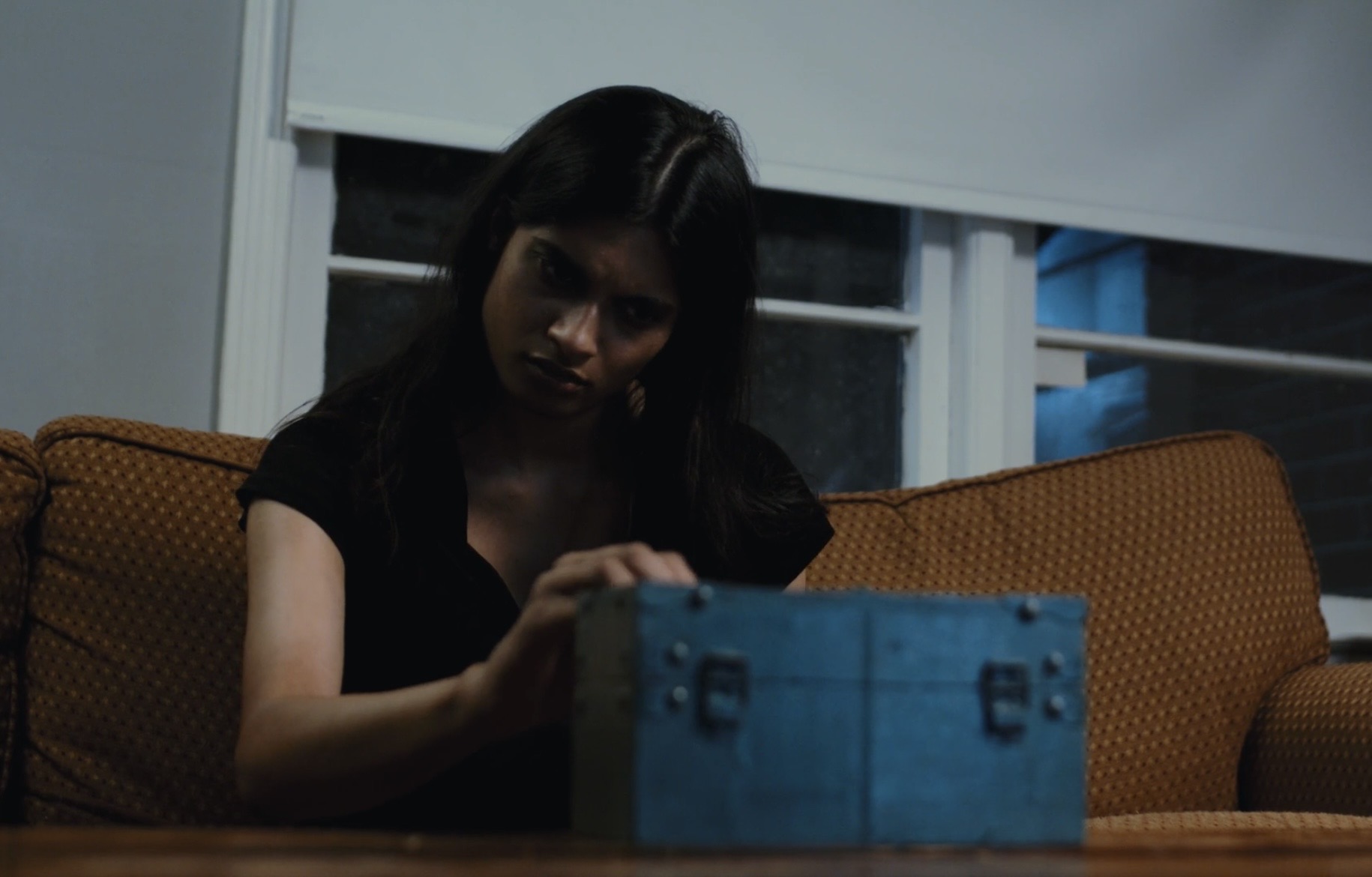 A woman with dark hair sits at a table in a dimly lit room, looking at a blue box.