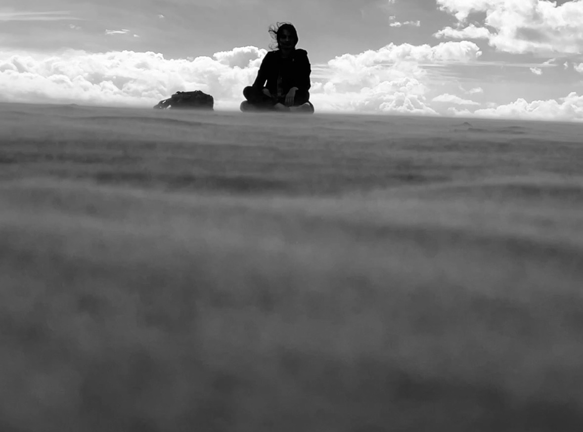 Person sitting on a beach with clouds in the sky, viewed from a low angle, black and white.