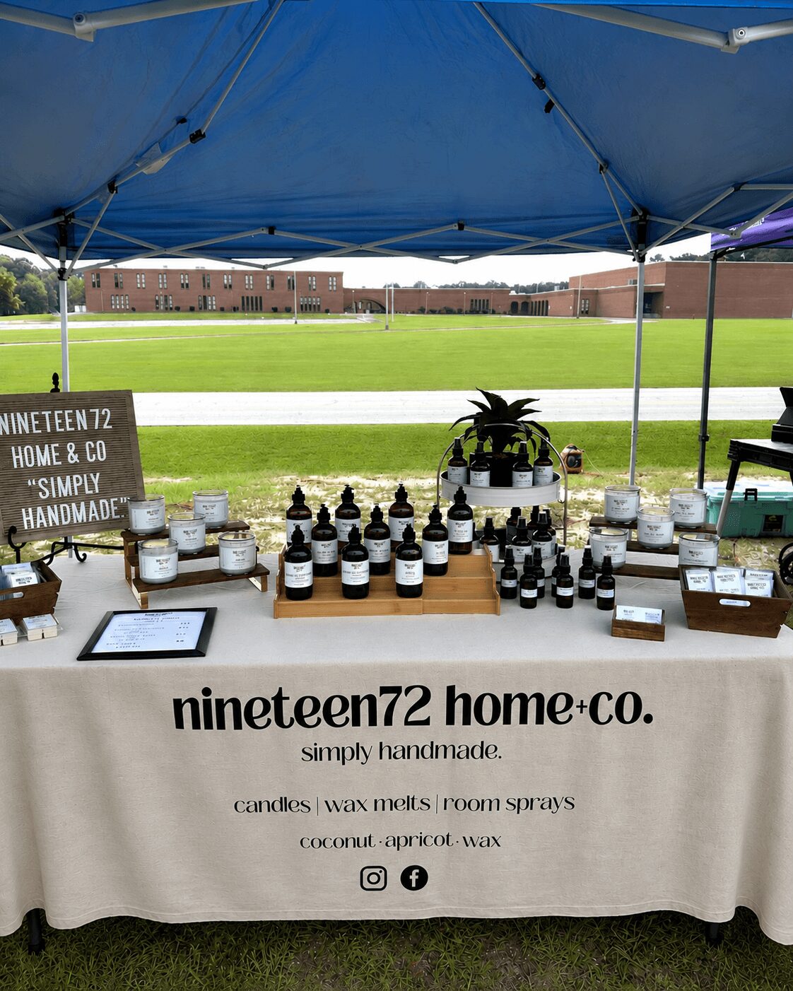 Table with handmade candles and sprays under a blue canopy, with a grassy field and brick buildings in the background.