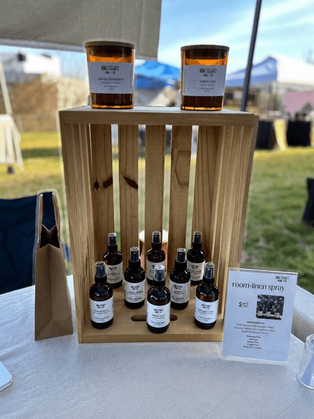 Wooden display with small bottles and two glasses of dark liquid outdoors, with tents and sky in background.