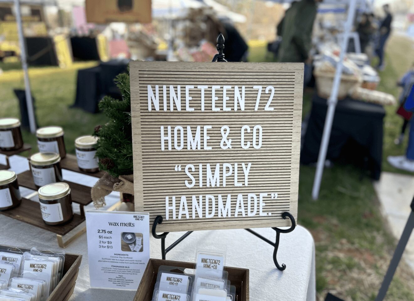 A sign reads 'NINETTEN 72 HOME & CO SIMPLY HANDMADE' at an outdoor market stall, with jars and products displayed nearby.