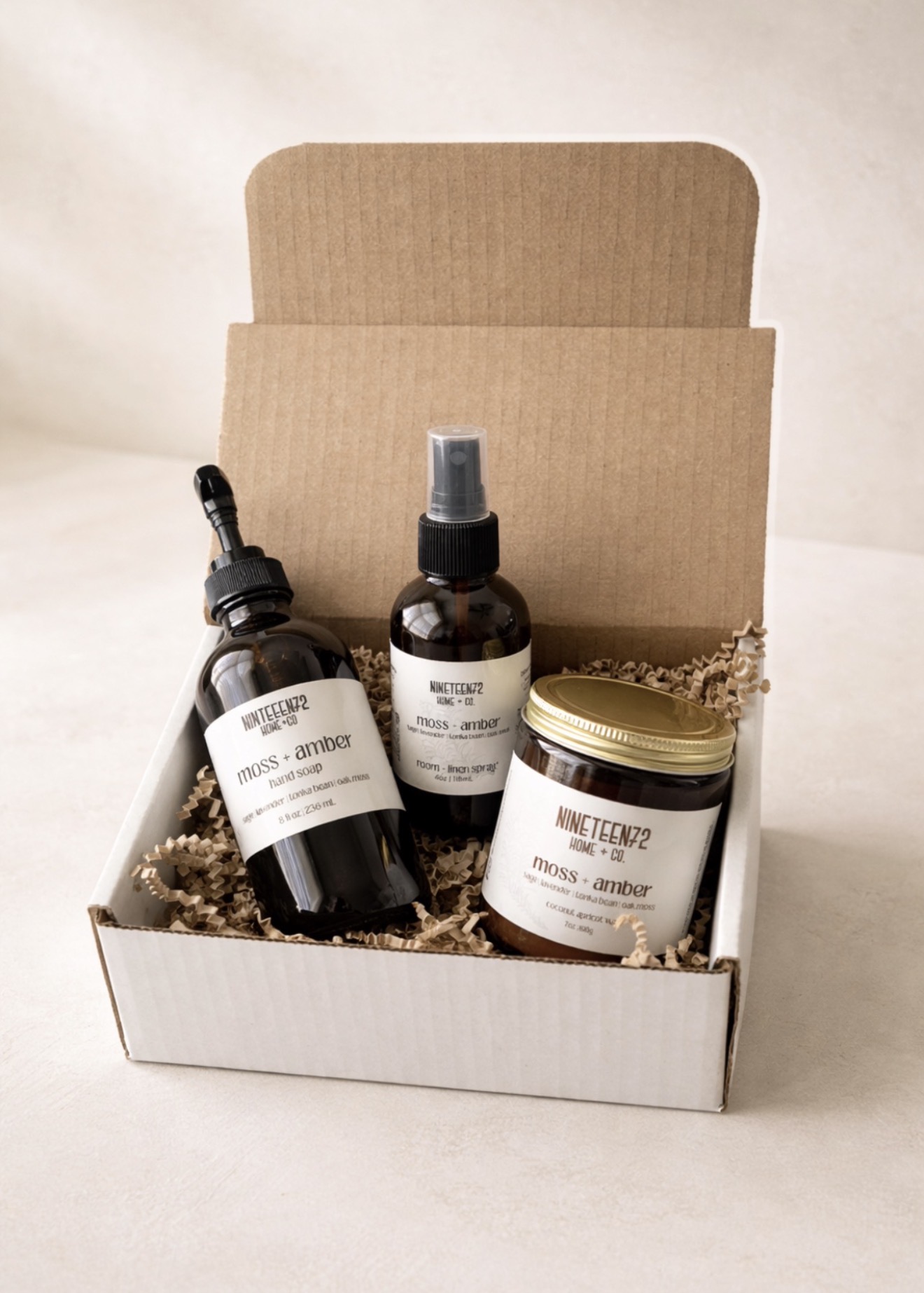 Open white box containing two dark bottles with spray tops and a jar with a gold lid, all on a bed of shredded paper.