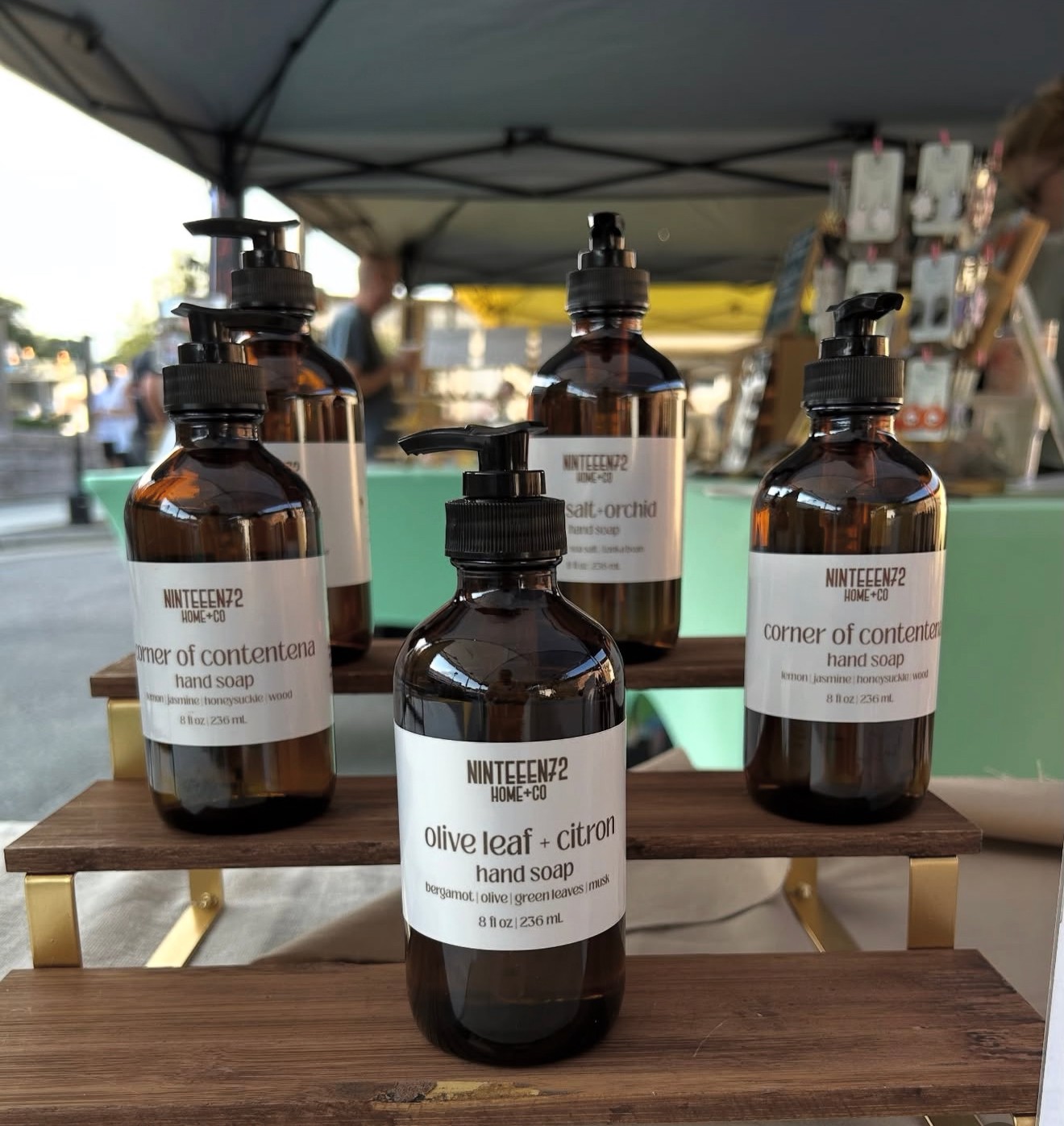 Five amber glass bottles with black pump dispensers on a wooden table, labels read 'olive leaf - citron hand soap'.