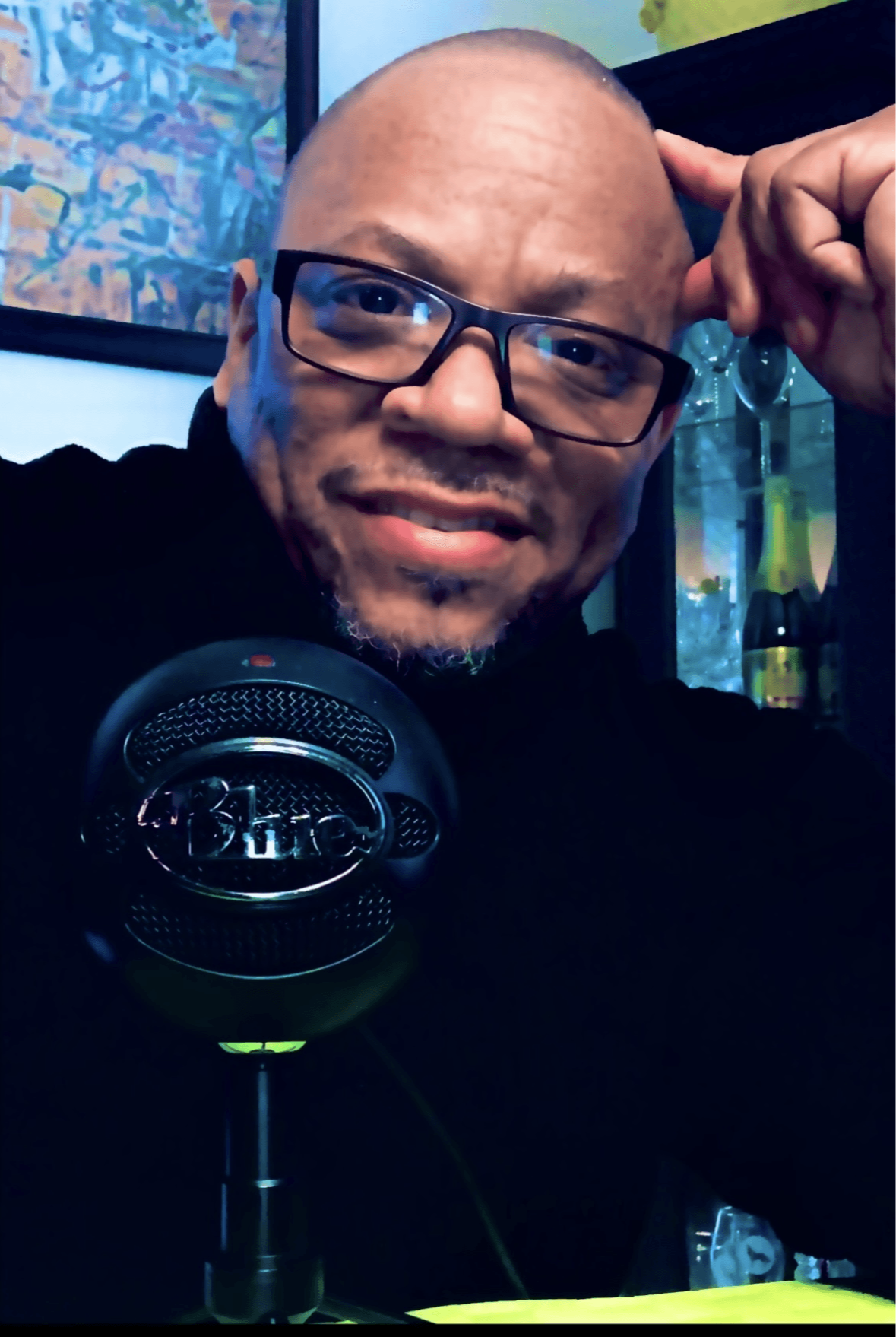 Man with glasses smiling, holding his head, in front of a microphone and colorful background.