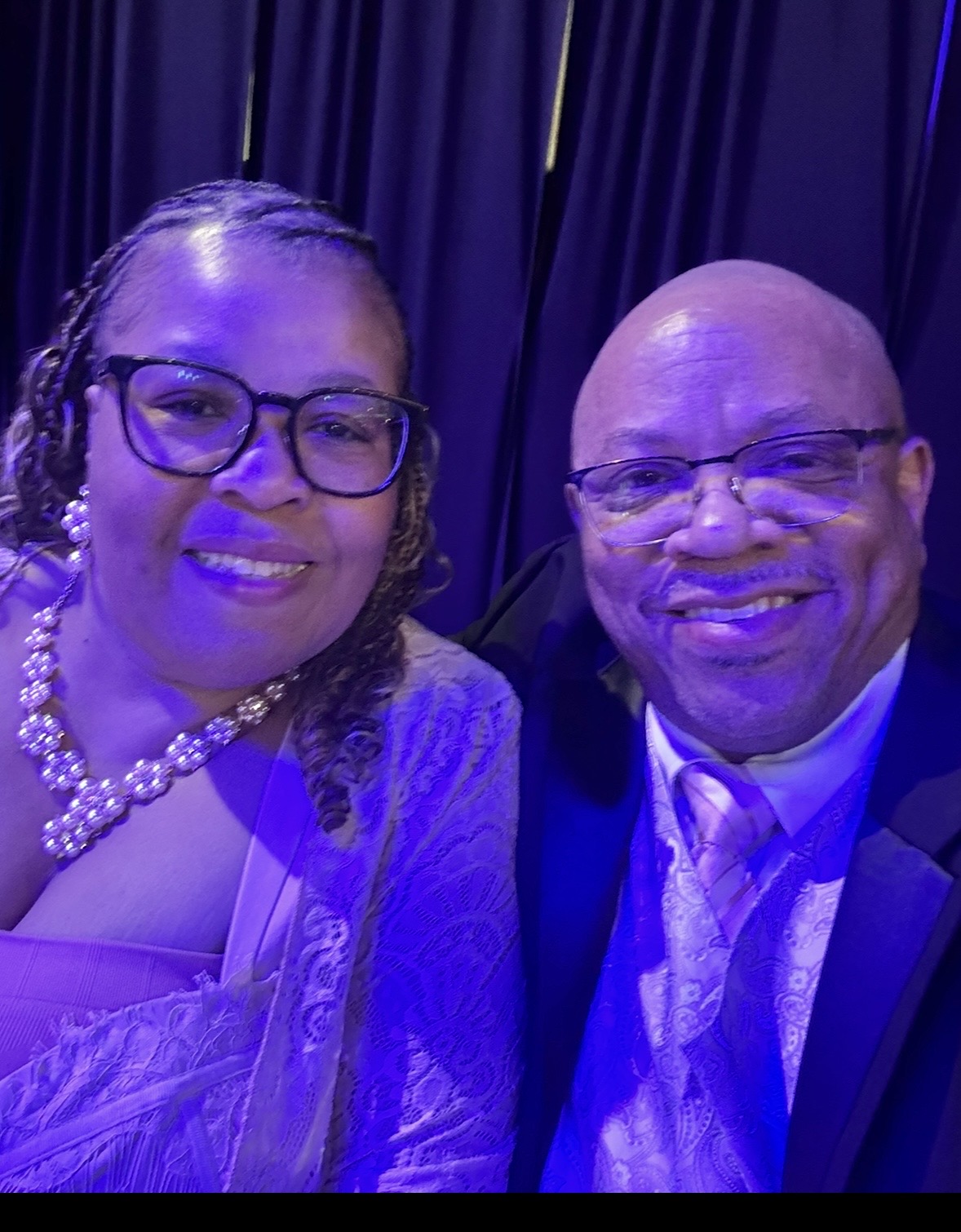 Two people smiling, one wearing glasses and a pearl necklace, the other bald, both dressed formally, with dark curtains in the background.