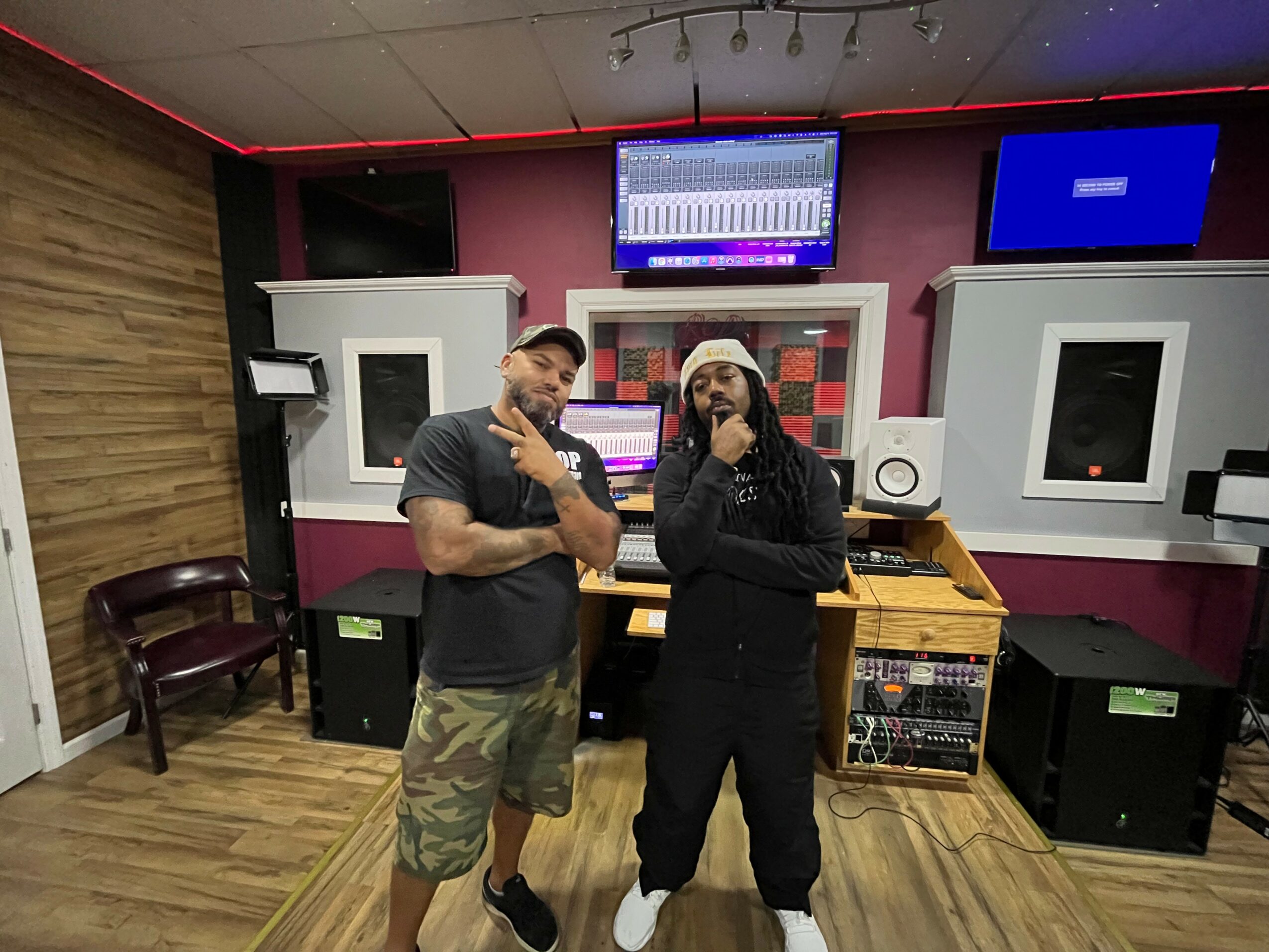 Two people stand in a music studio with equipment, monitors, and a pink and white wall behind them.