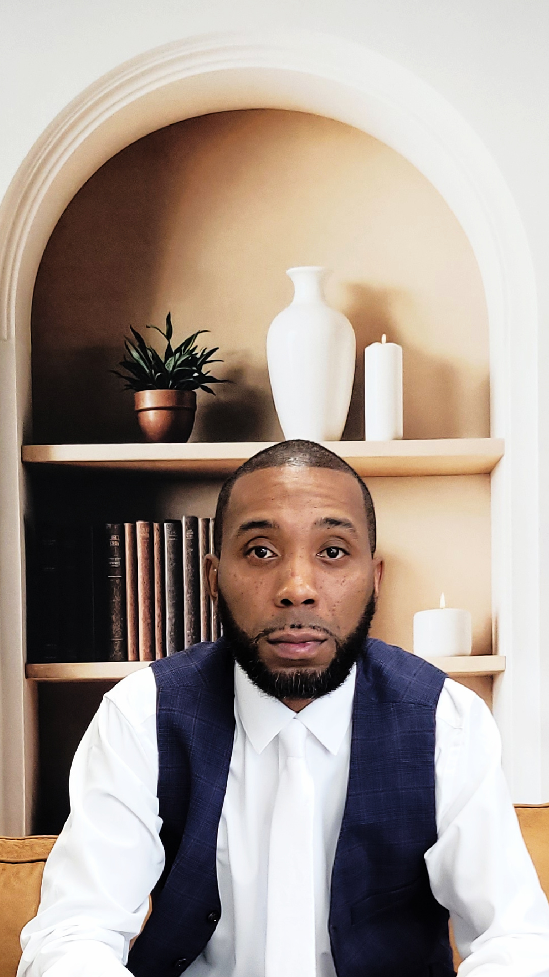 Man with short hair and beard wearing a white shirt and dark vest, sitting indoors with shelves and decorative items behind him.