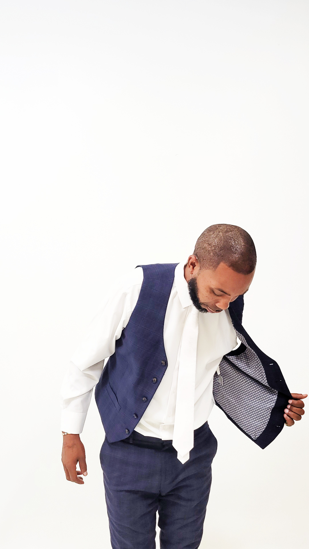 Man looking down, wearing a navy vest and pants, holding open a jacket to show its interior, against a white background.