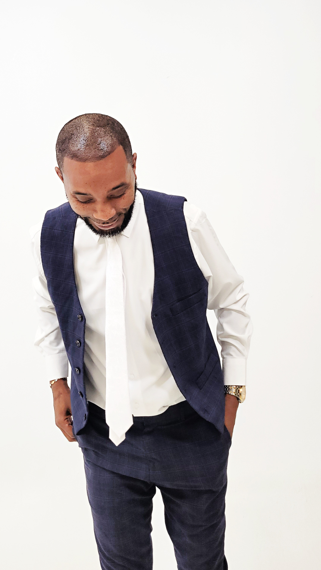 Man with short hair and beard wearing a white shirt, dark vest, and pants, looking down with hands in pockets.