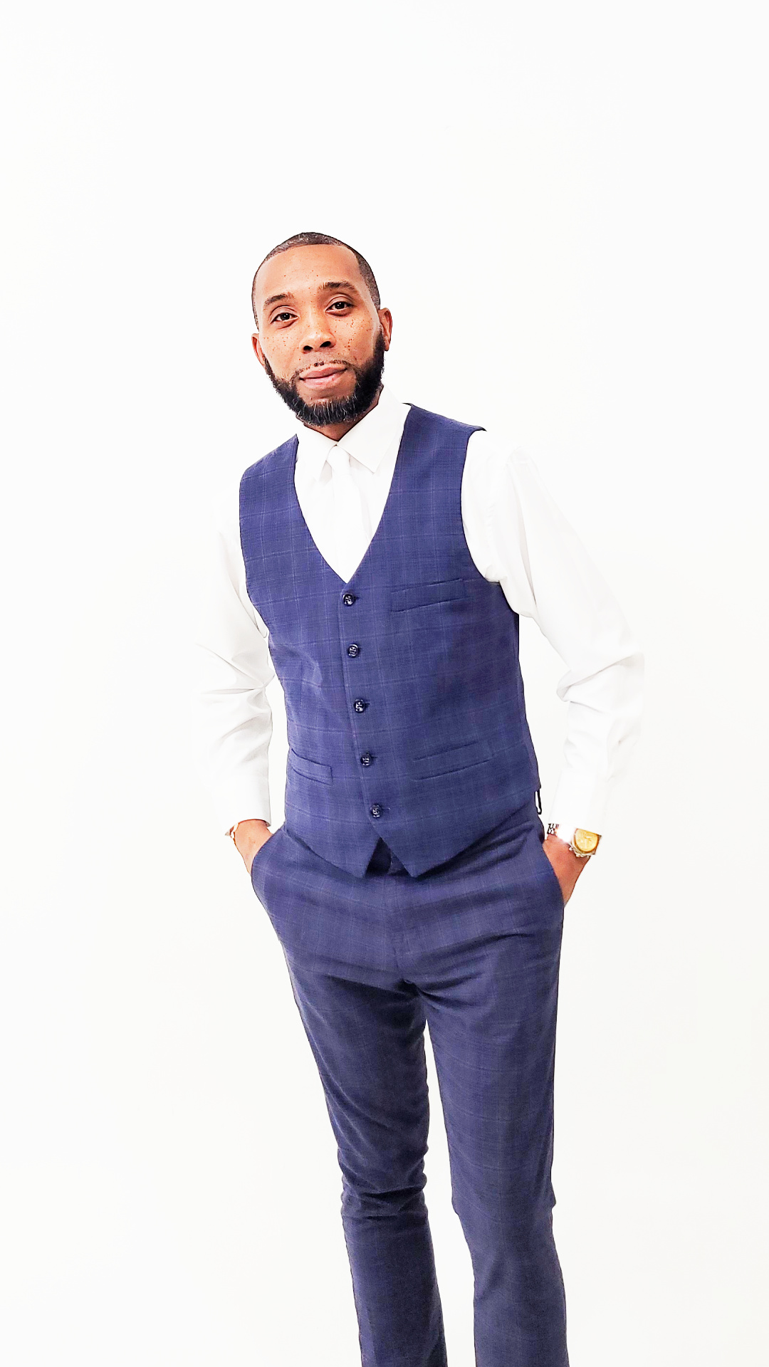 Man in a blue vest and pants with a white shirt, standing with hands in pockets, smiling, against a white background.