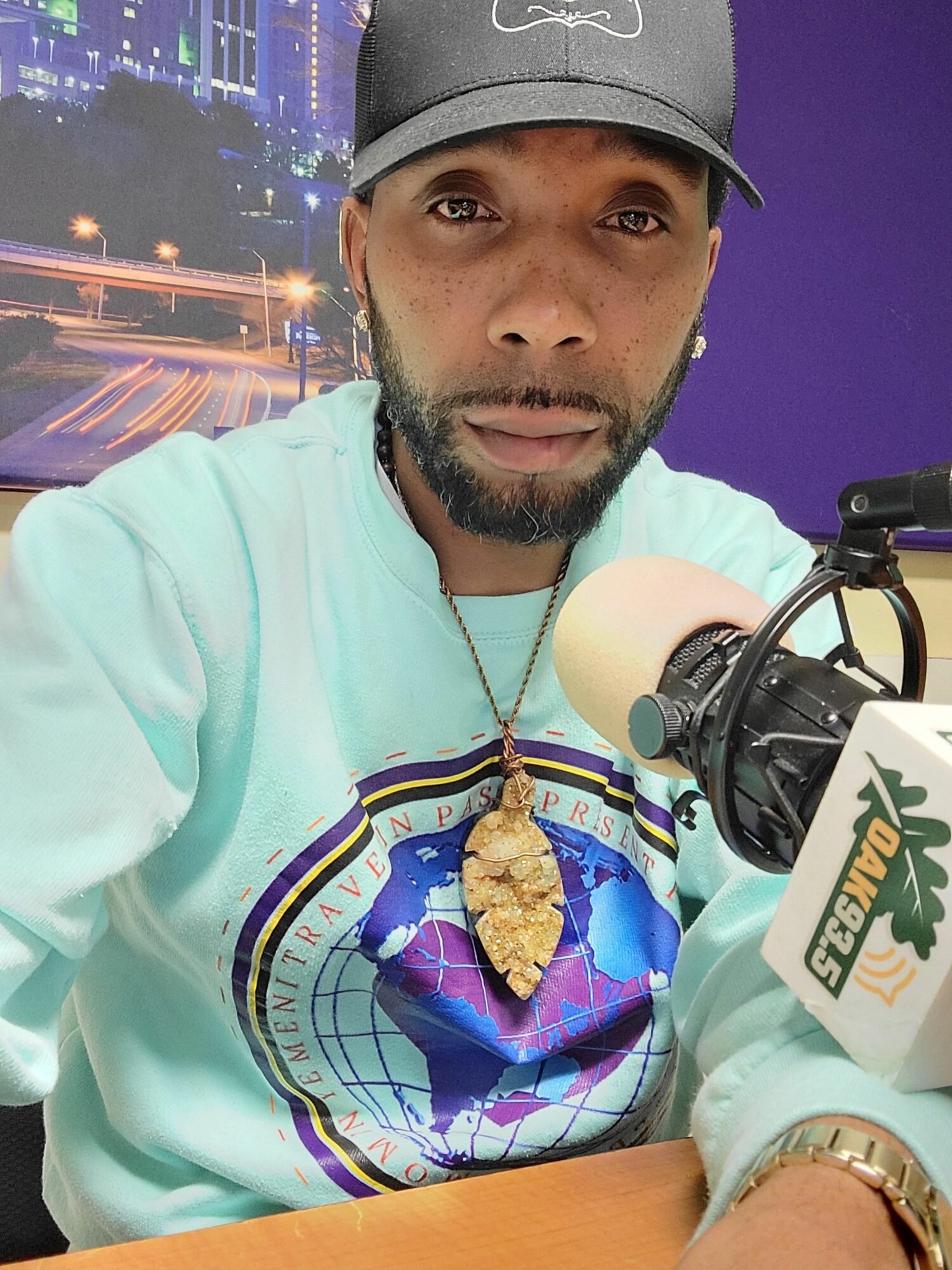 Man wearing a cap and hoodie, sitting at a desk with a microphone, cityscape background, looking at camera.